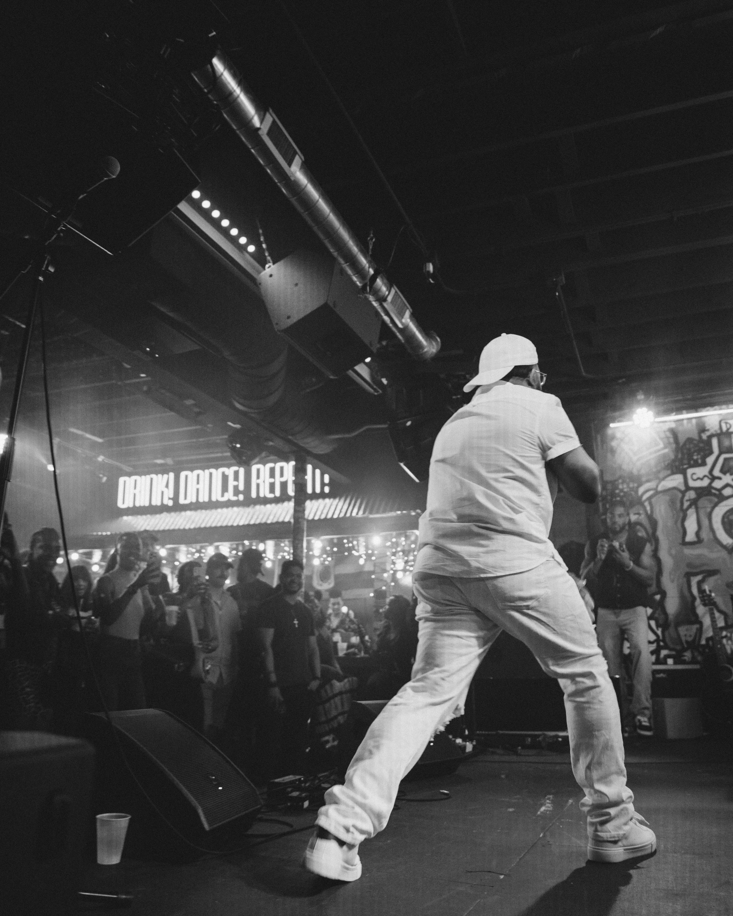 A person in a white hat and white clothing dancing on stage with a crowd in the background, illuminated by stage lights, in a dark room with graffiti on the walls.