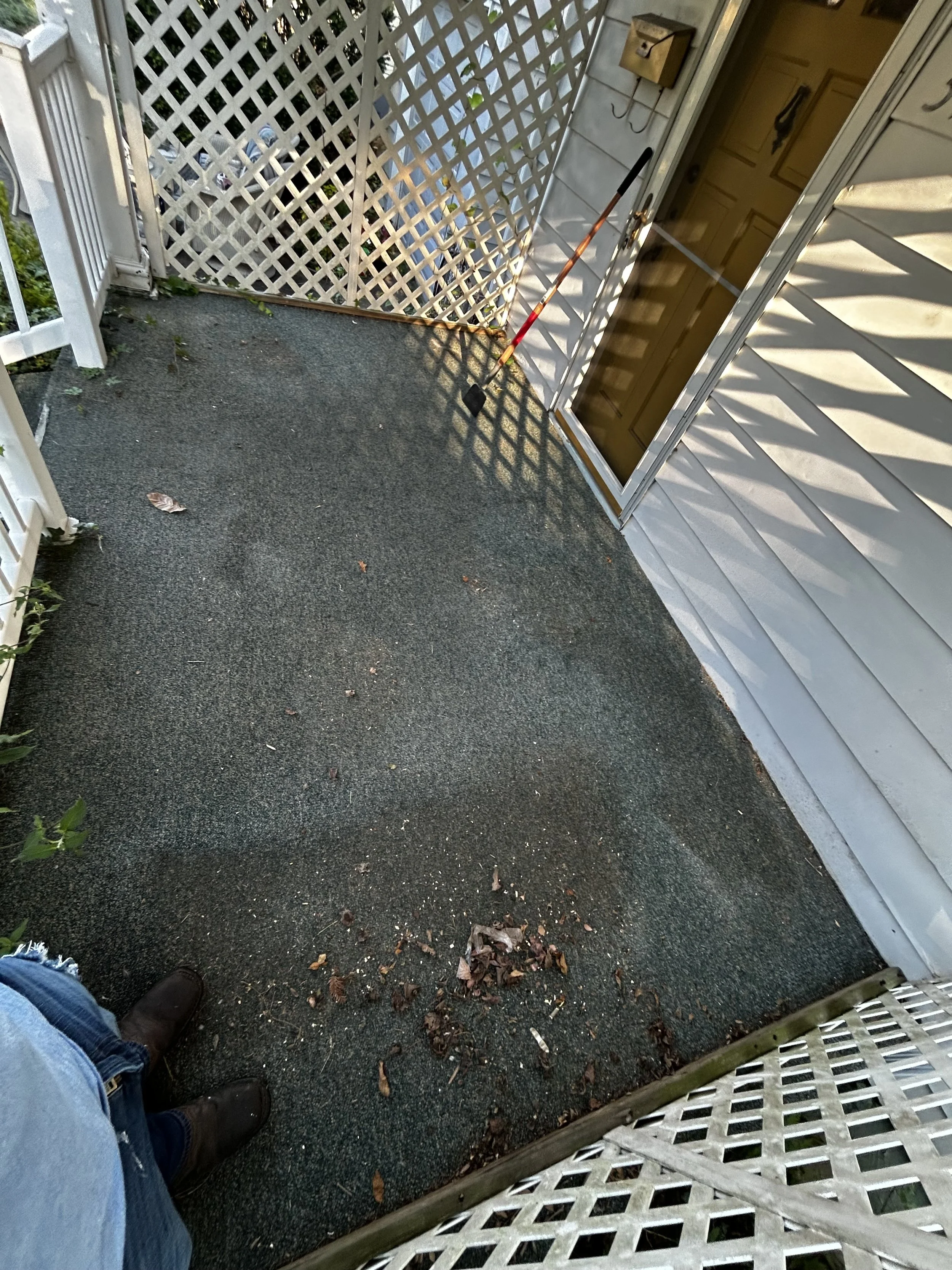 View of a porch with a black carpeted floor, a person wearing blue jeans and black boots standing in the lower left corner, a white lattice fence in the background, a yellow door with a black hook, a brass doorknob, and a mail slot, a yellow mailbox mounted on the wall, a gardening tool with a long handle and black head leaning against the door, dry leaves scattered on the floor, sunlight casting shadows, and a part of a white railing on the left side.