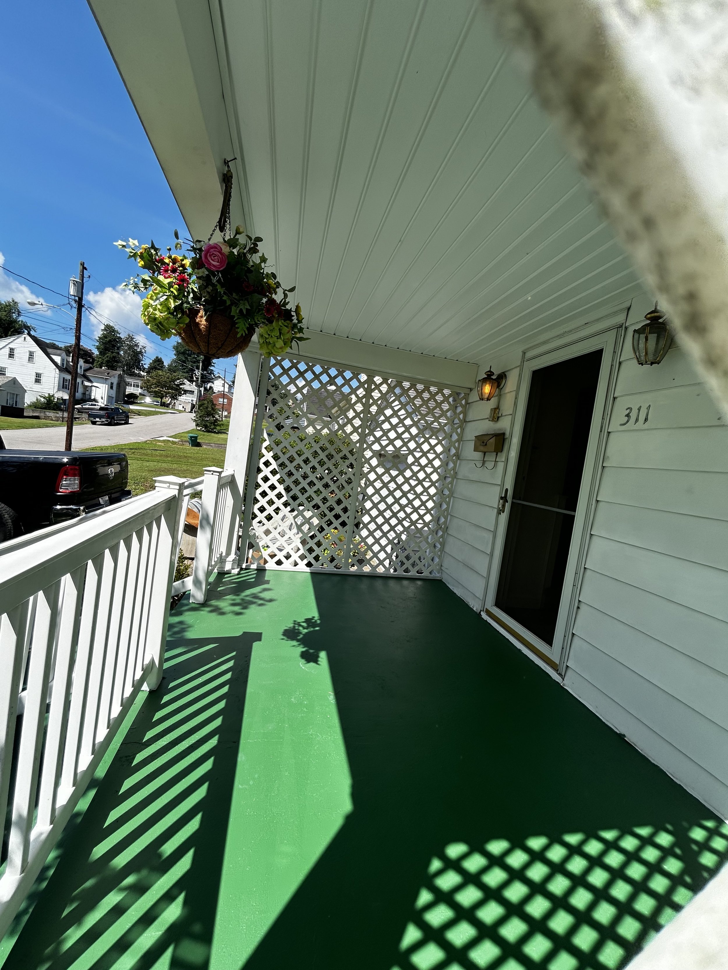 Front porch of a house with a bright green floor, white railing, hanging flower basket, and lattice privacy screen, with a view of a neighborhood street with cars and houses in the background.