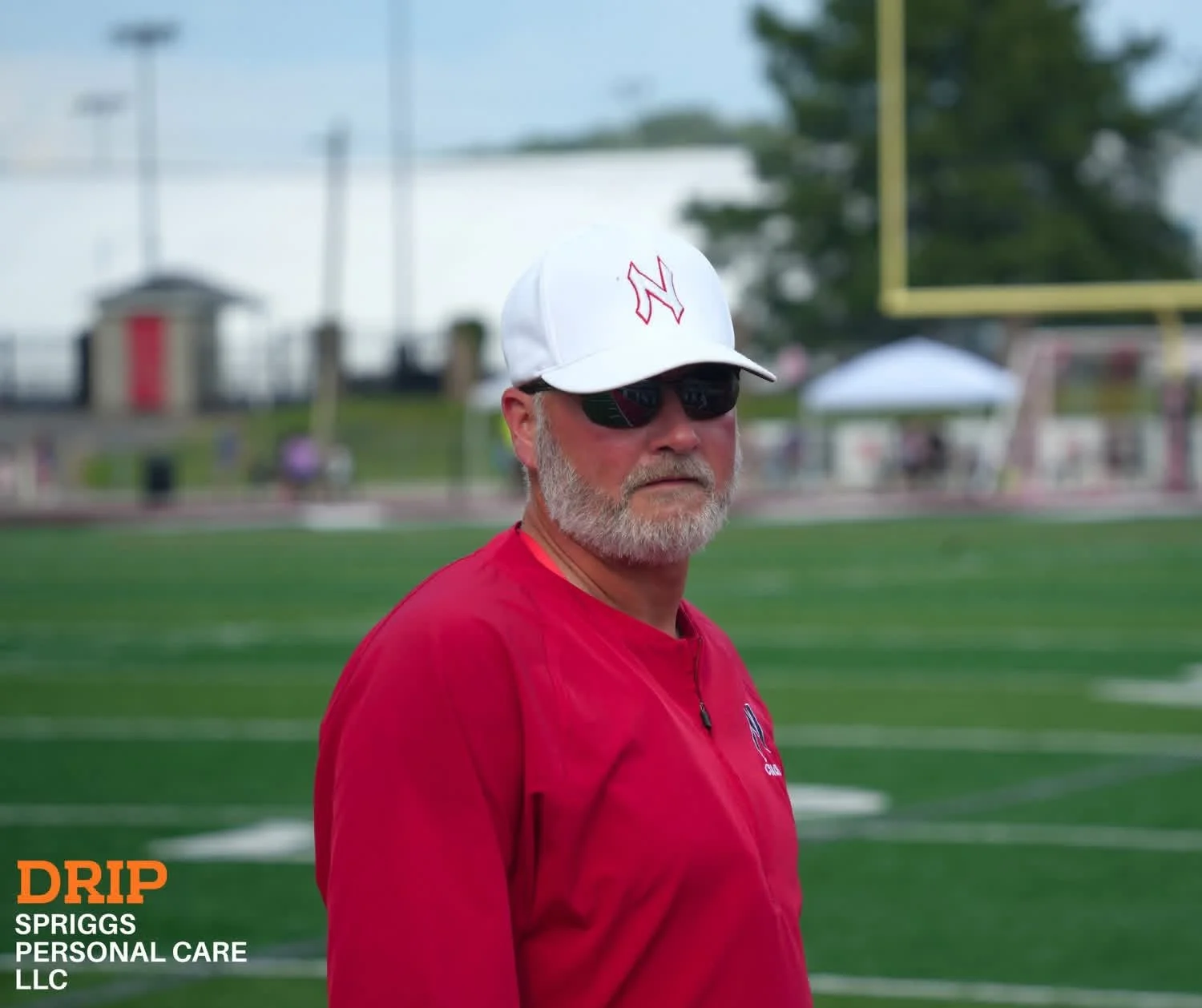 A man wearing a white cap, black sunglasses, and a red jacket stands on a sports field. The background shows a football goalpost, trees, and some umbrellas, indicating an outdoor athletic setting.