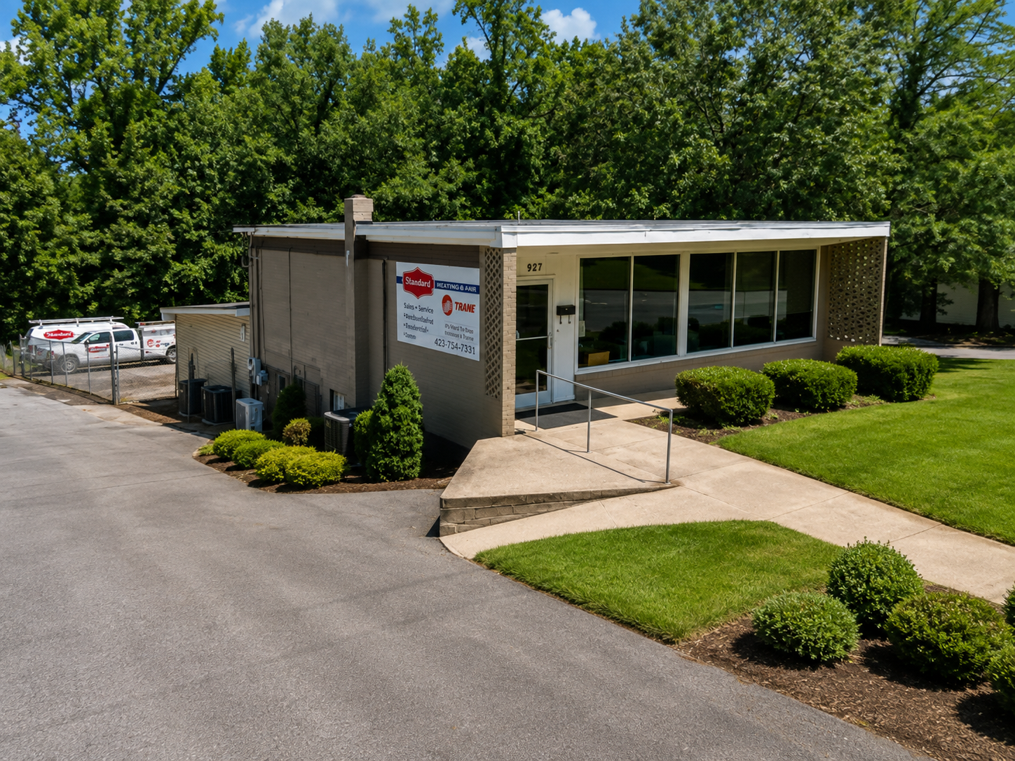 Office building with large front windows, surrounded by green shrubs and a well-maintained lawn, with a sign indicating HVAC services and vehicles parked nearby.