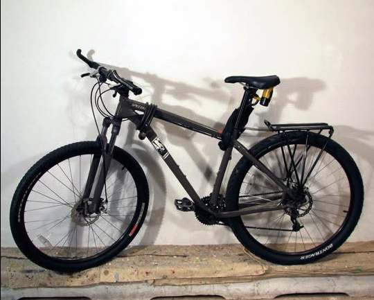 Black mountain bike with front suspension and rear rack, leaning against a plain white wall.