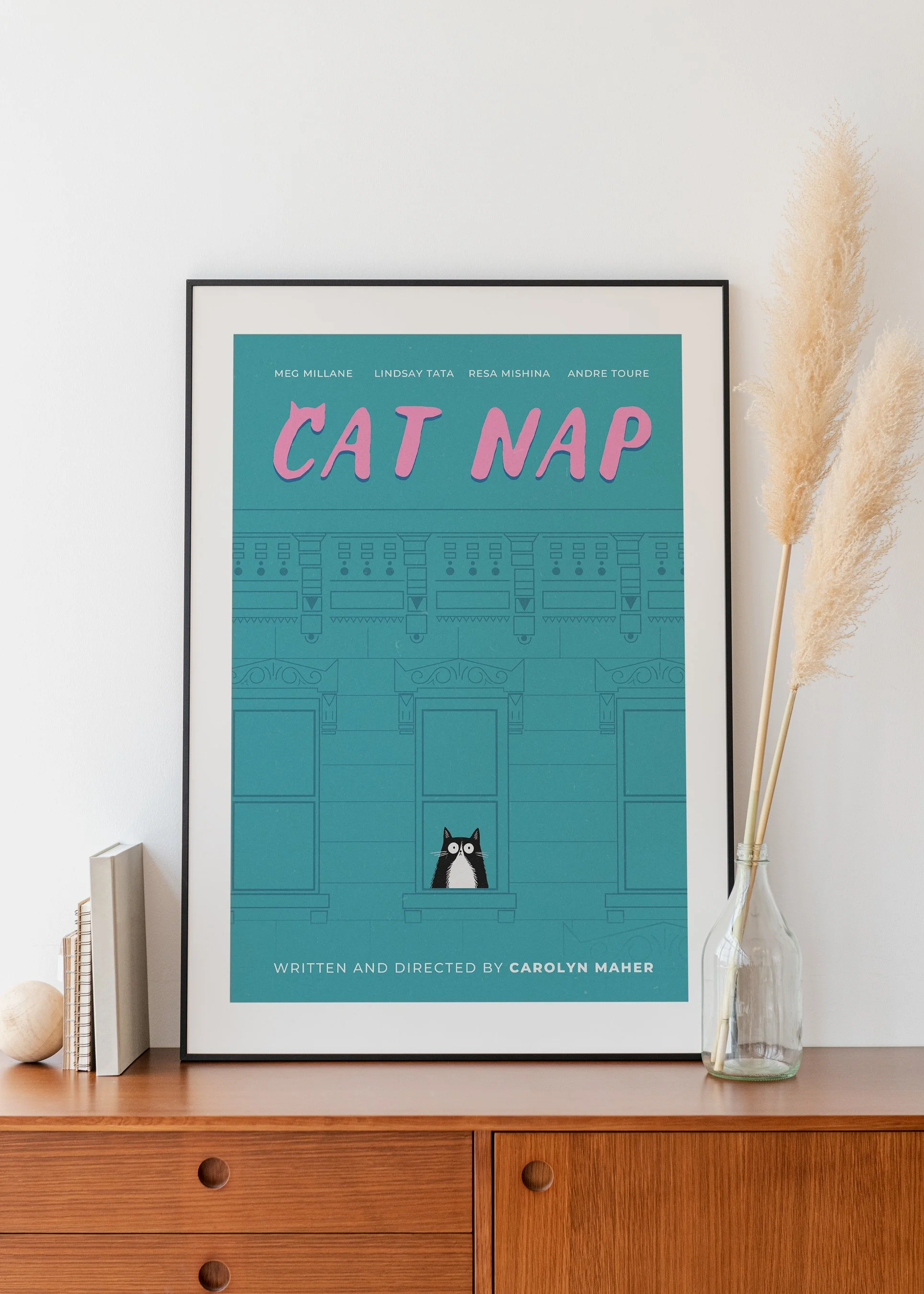 Close-up of a framed poster on a wooden dresser, featuring the title 'Cat Nap' in large pink letters, a small illustration of a black and white cat, and text indicating it is written and directed by Carolyn Maher. To the right of the poster, a glass vase holds three dried pampas grass stems. A couple of books and a light-colored spherical object are on the left side of the dresser.