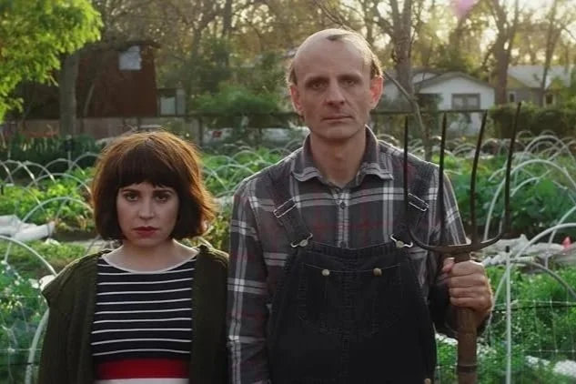 A man and woman standing in a garden with green plants and arched supports, against a backdrop of trees and houses, during sunlight.