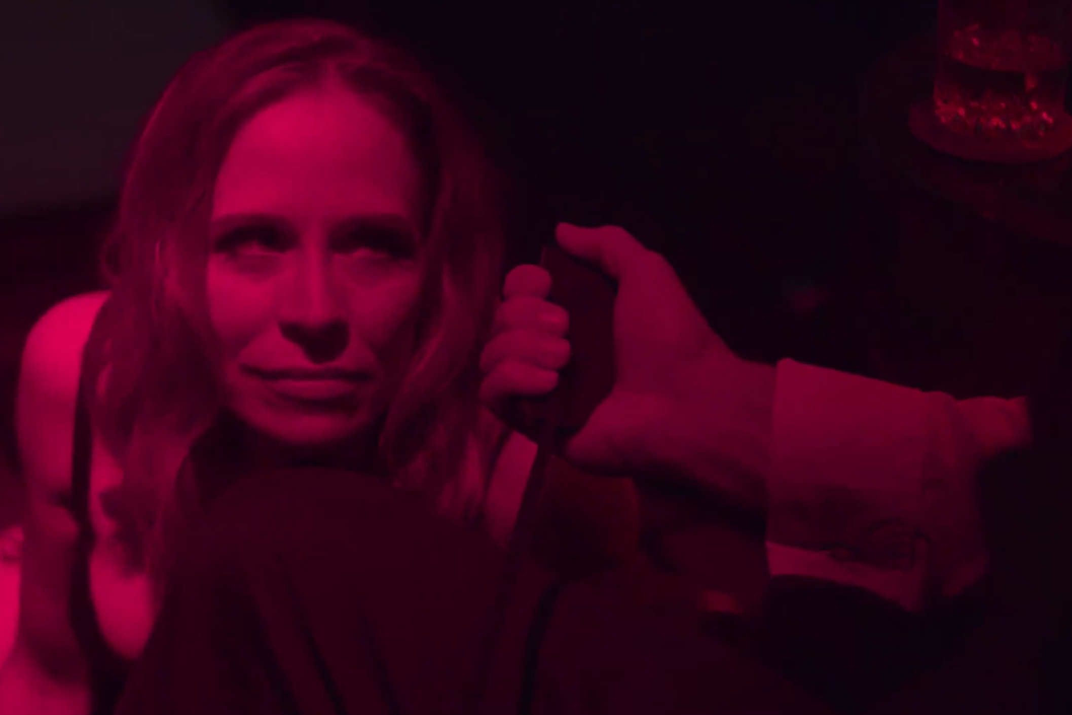 A woman in a dark room with pink lighting, with her head tilted slightly, smiling softly; a hand moves towards her ear, adjusting her hair.
