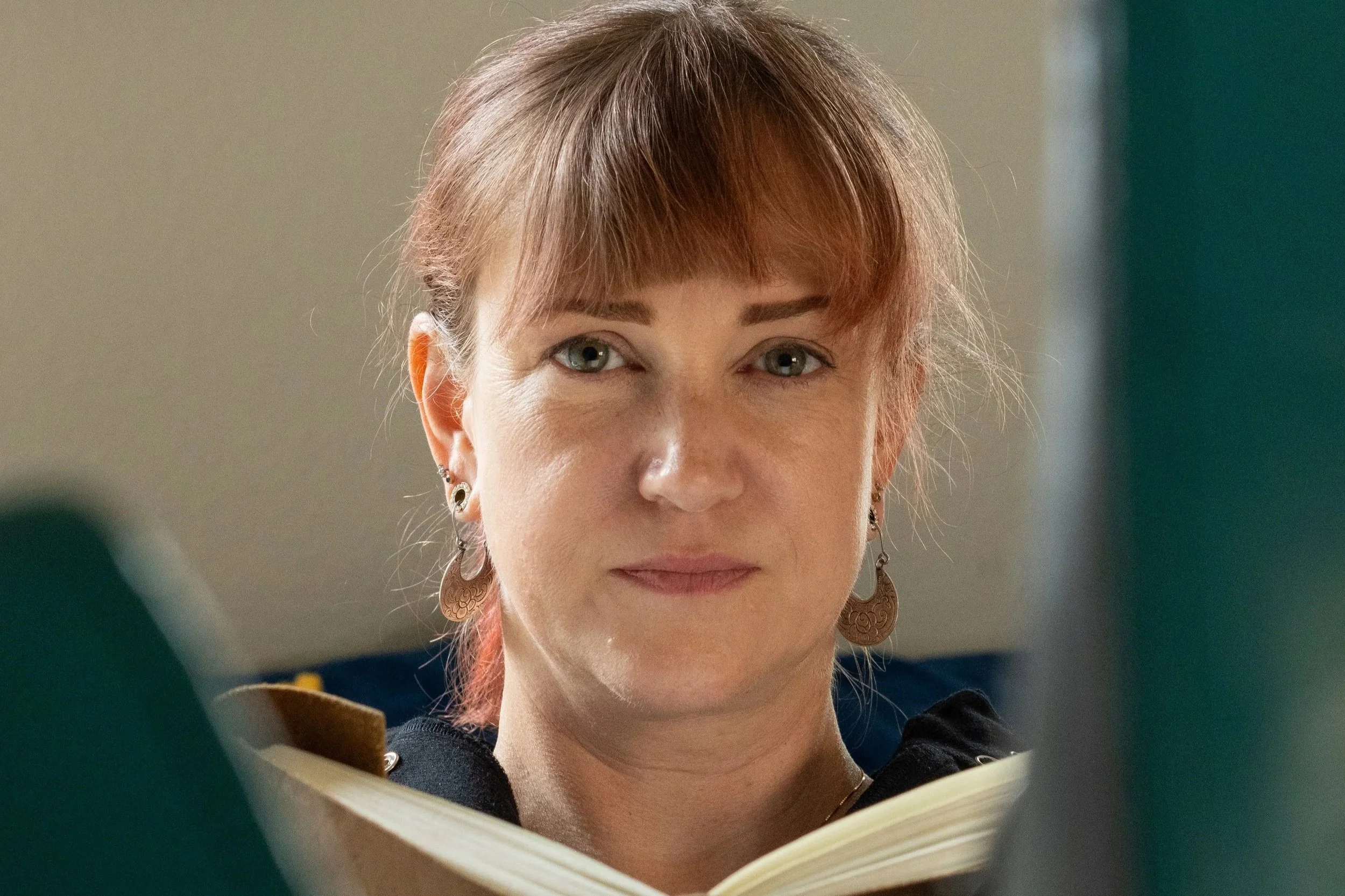 A woman with short reddish hair and wearing earrings looks at a mirror or a camera while reading a book or a tablet.