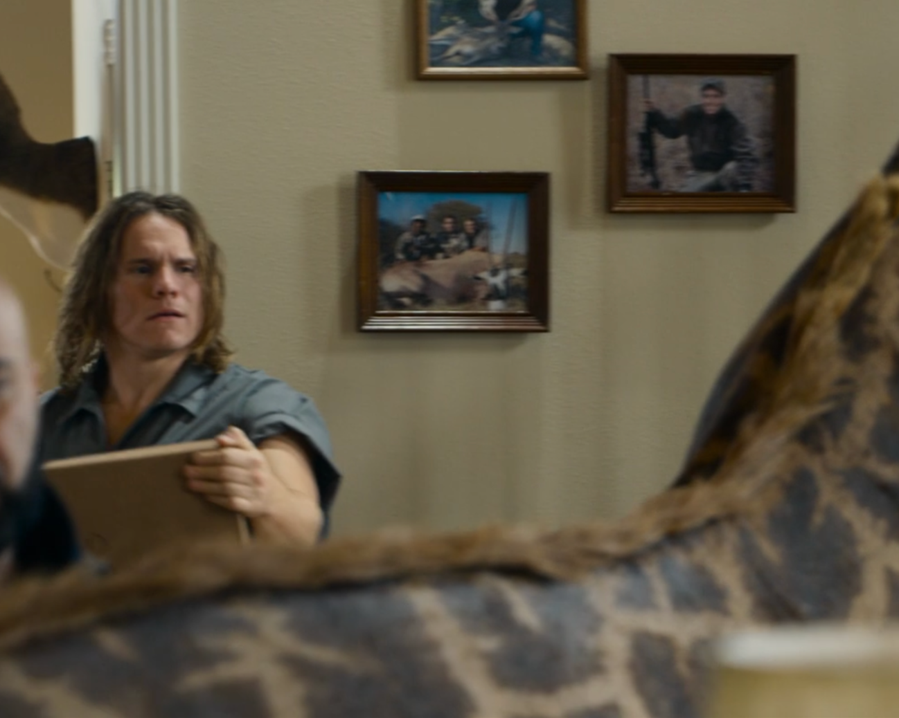 A scene from a movie or TV show featuring a man with shoulder-length hair holding a clipboard, looking serious or concerned. The background shows a wall with three framed photographs, including one of a man sitting outdoors and another of two people with a large animal. Part of a person's body with animal print clothing is visible in the foreground.