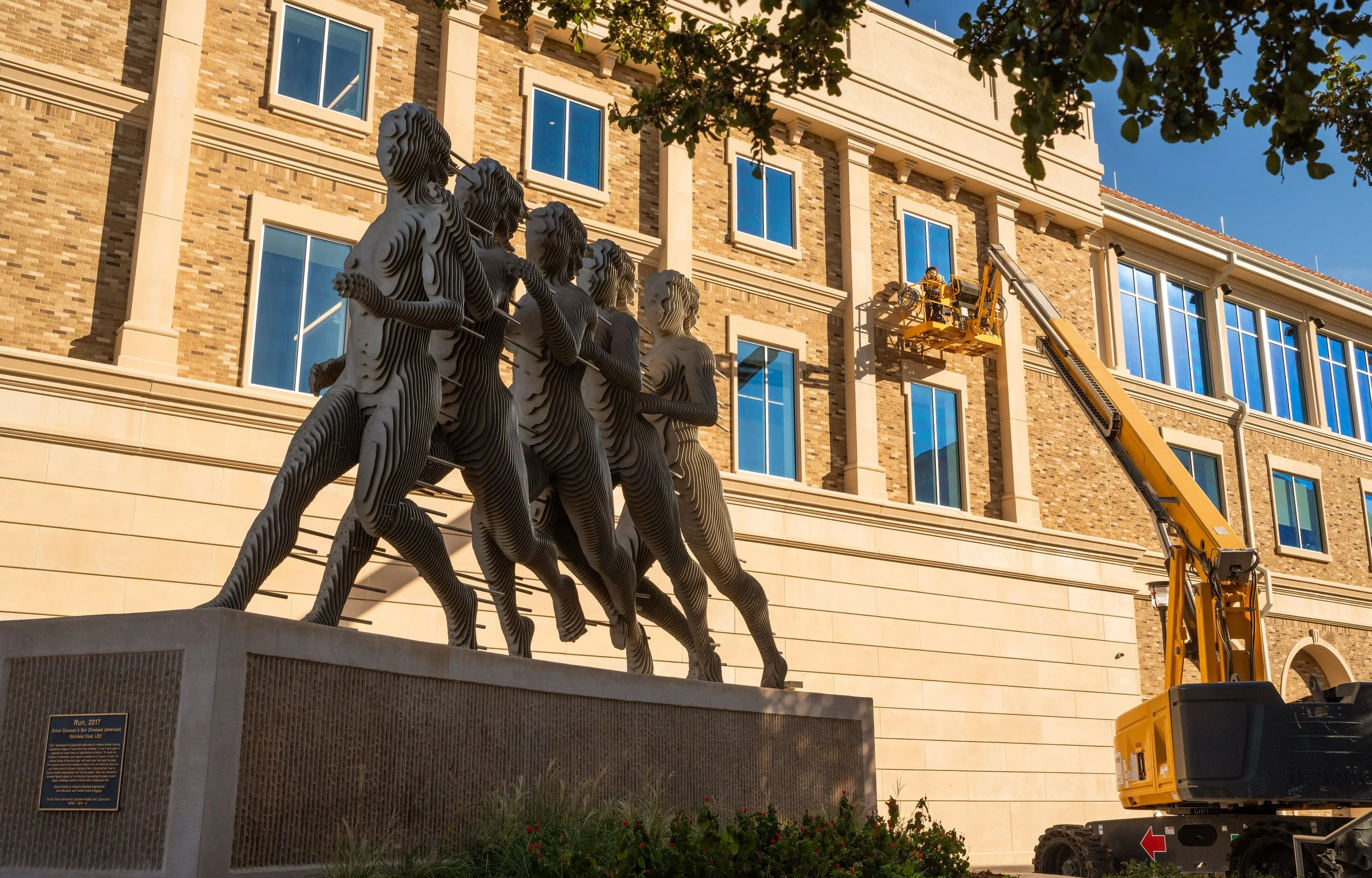 A large bronze sculpture of people running in a line, holding sticks in front of a brick building with blue windows. A construction lift is parked nearby with a worker on it, installing glass on the building facade.