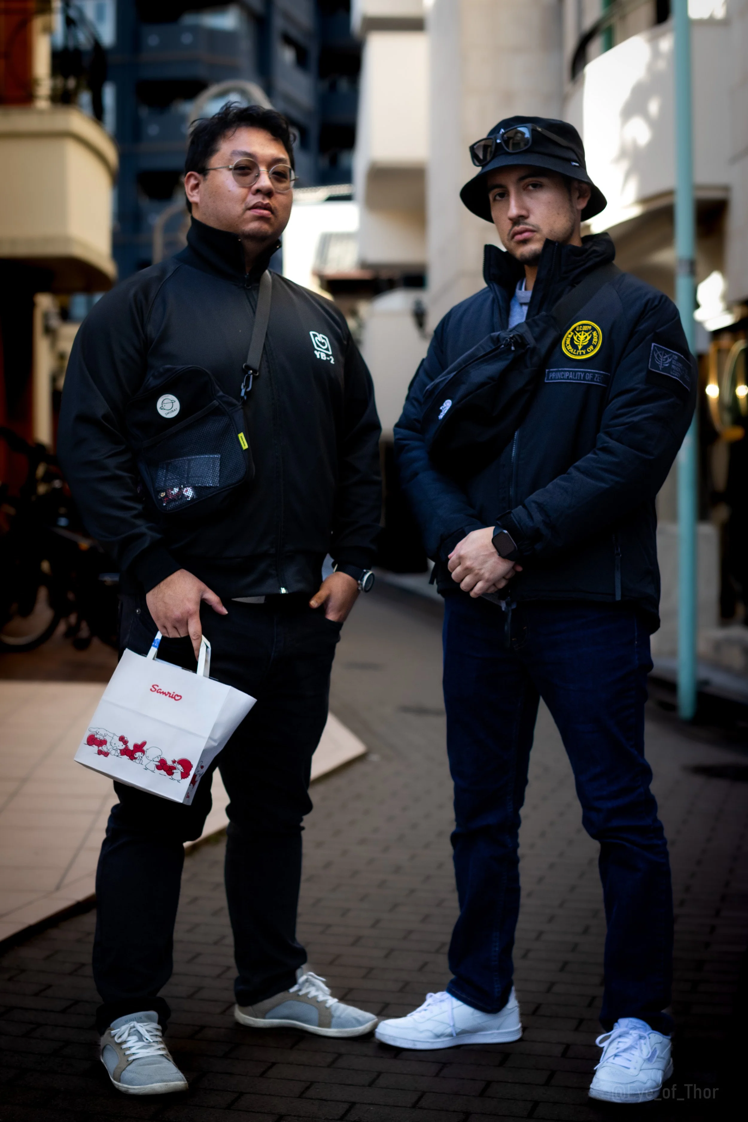 Two men standing outdoors in casual clothing, posing for a photo, with buildings and a street background.