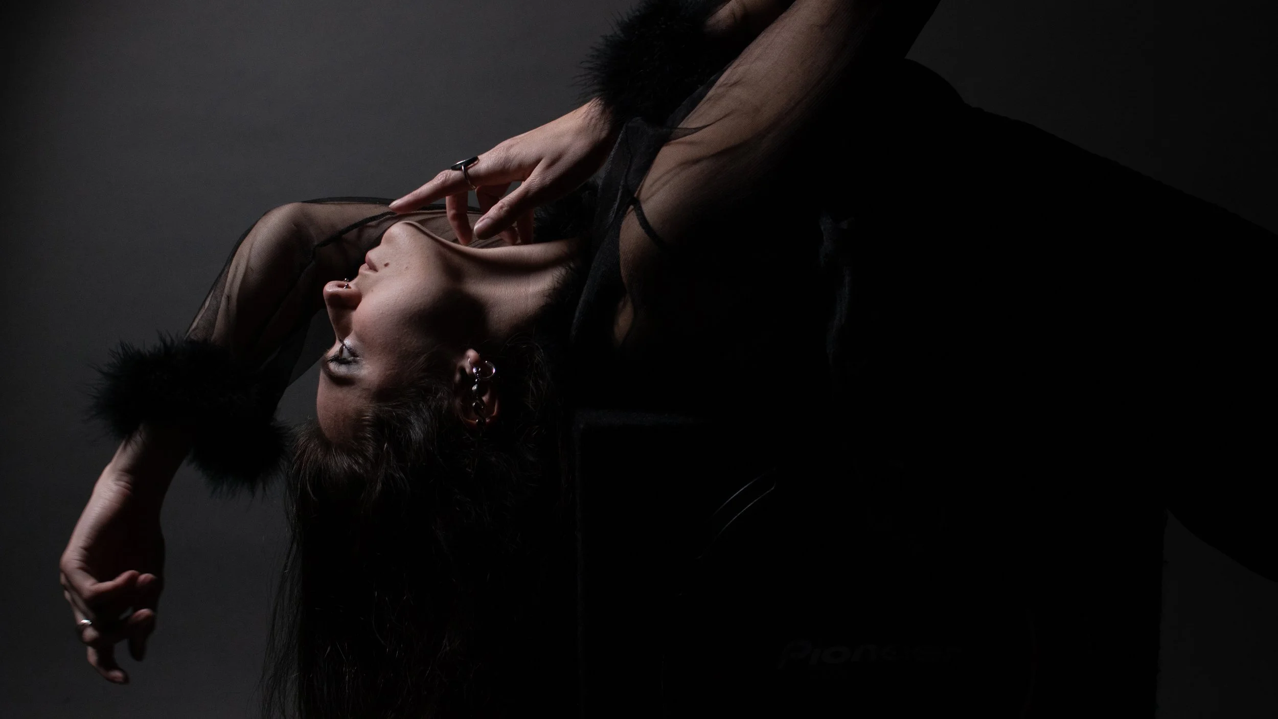 Person with dark hair wearing a black sheer top and jewelry, posing with hands near face and eyes closed against a dark background.