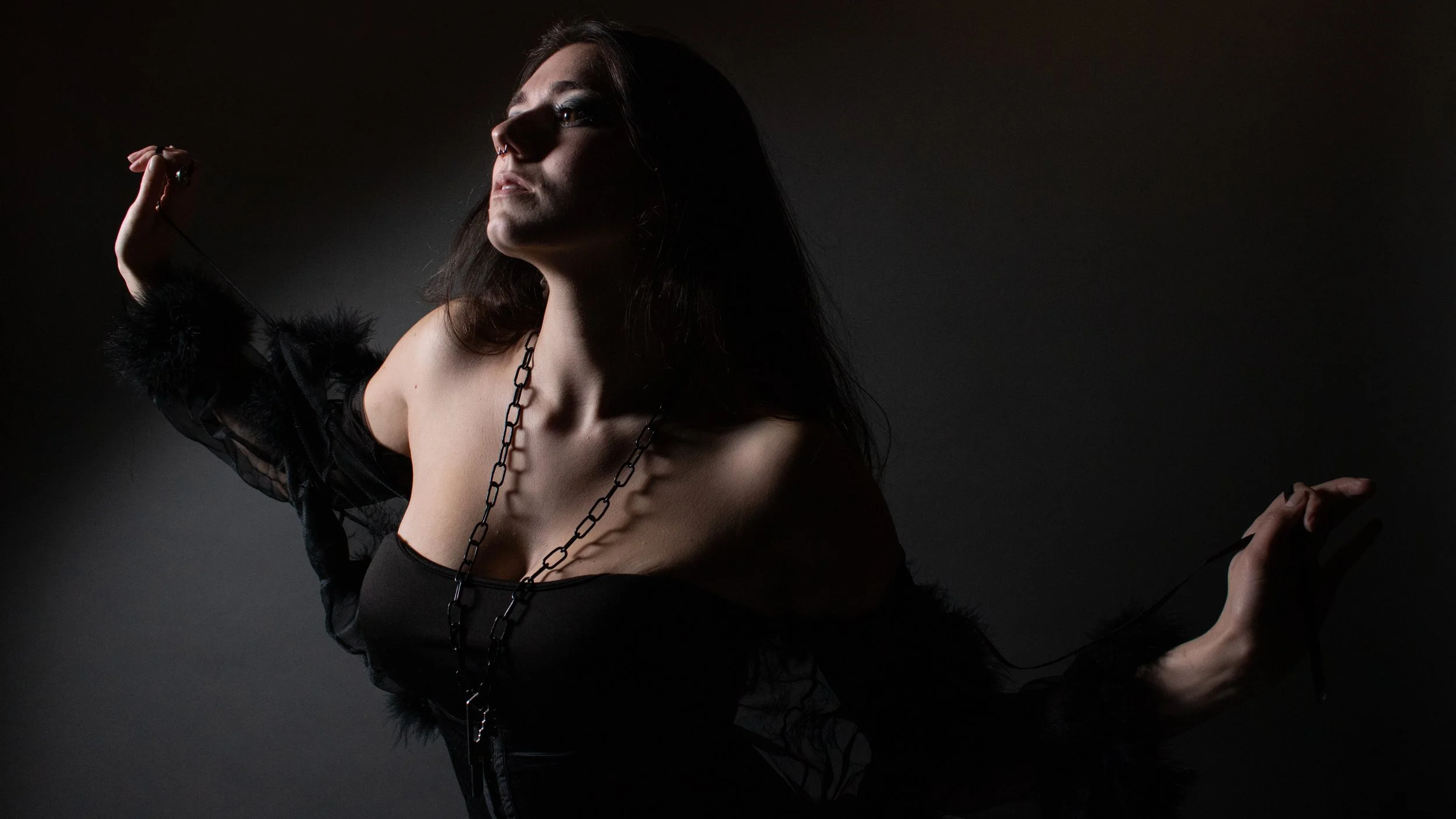 A woman with dark hair and light skin in dark clothing, wearing chunky chain necklaces, poses against a black background with dramatic lighting.