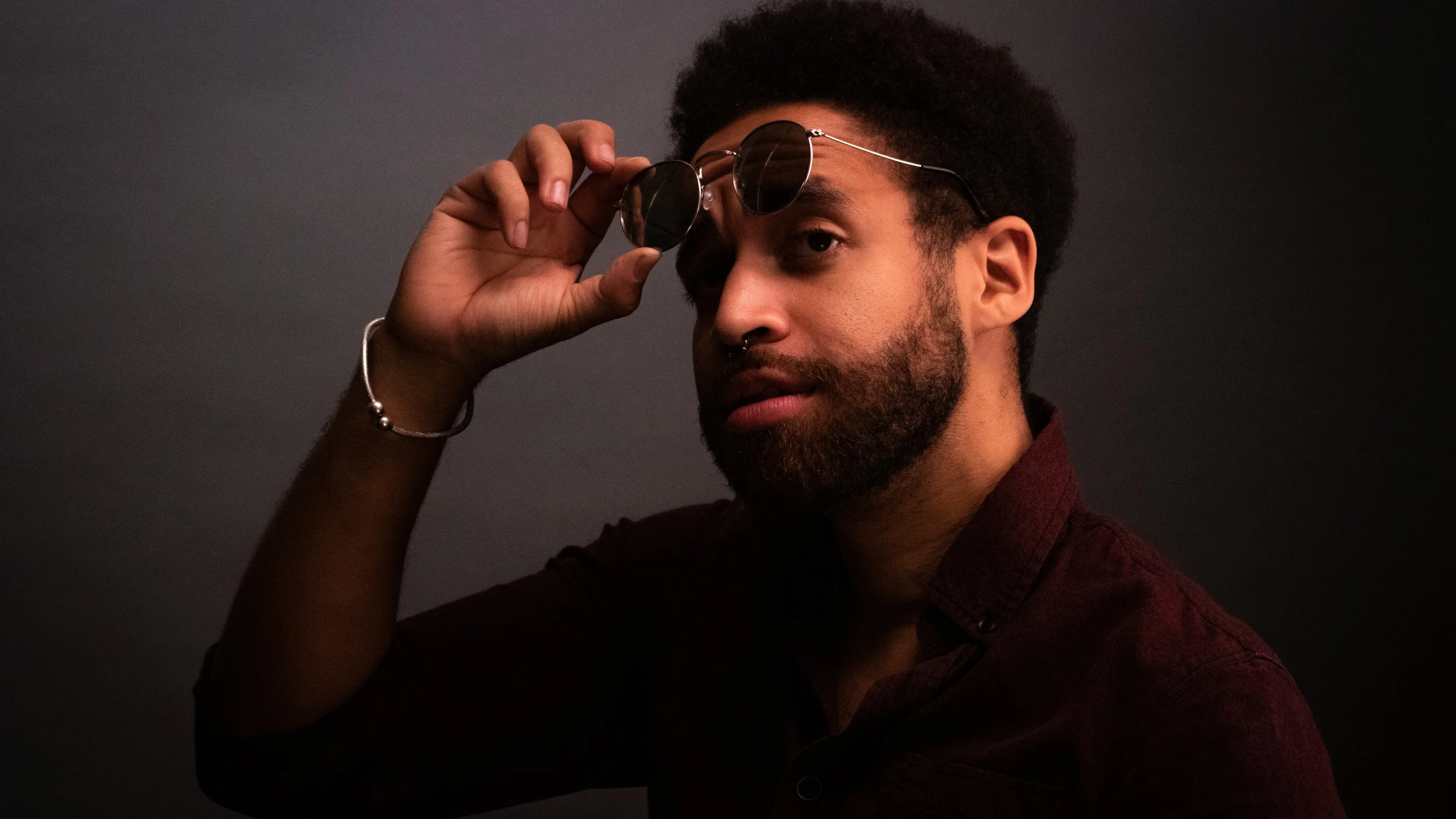 A young man with dark hair and a beard, wearing sunglasses and a dark red shirt, adjusts his sunglasses against a dark background.