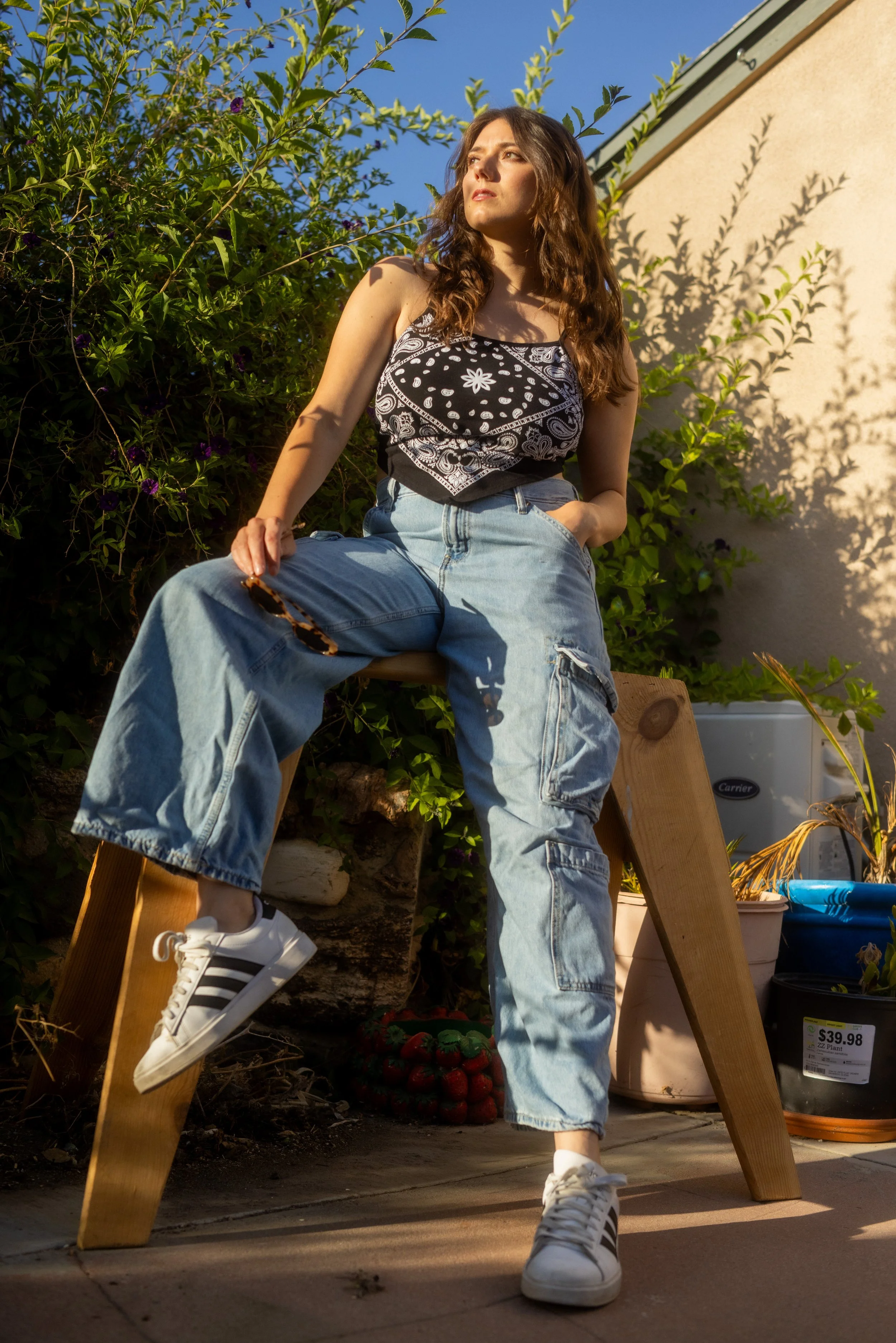 Young woman with wavy brown hair, wearing a black and white patterned tank top, ripped jeans, and white sneakers with black stripes, sitting on a wooden bench outdoors with green foliage and clear blue sky in the background.