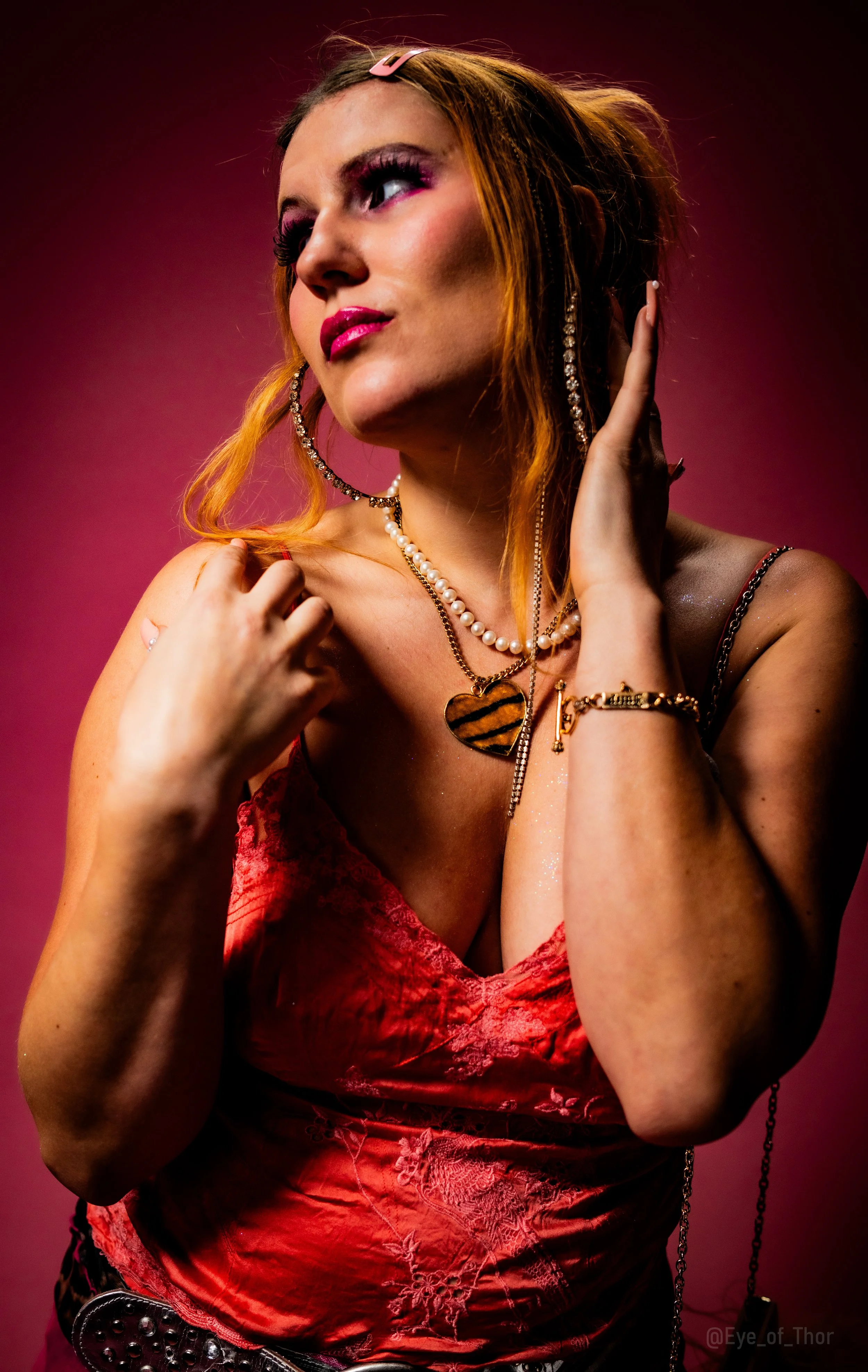 A woman with curly red hair wearing a red lace dress, jewelry including pearls and gold accessories, and makeup with pink lipstick and purple eyeshadow, posed against a dark pink background.