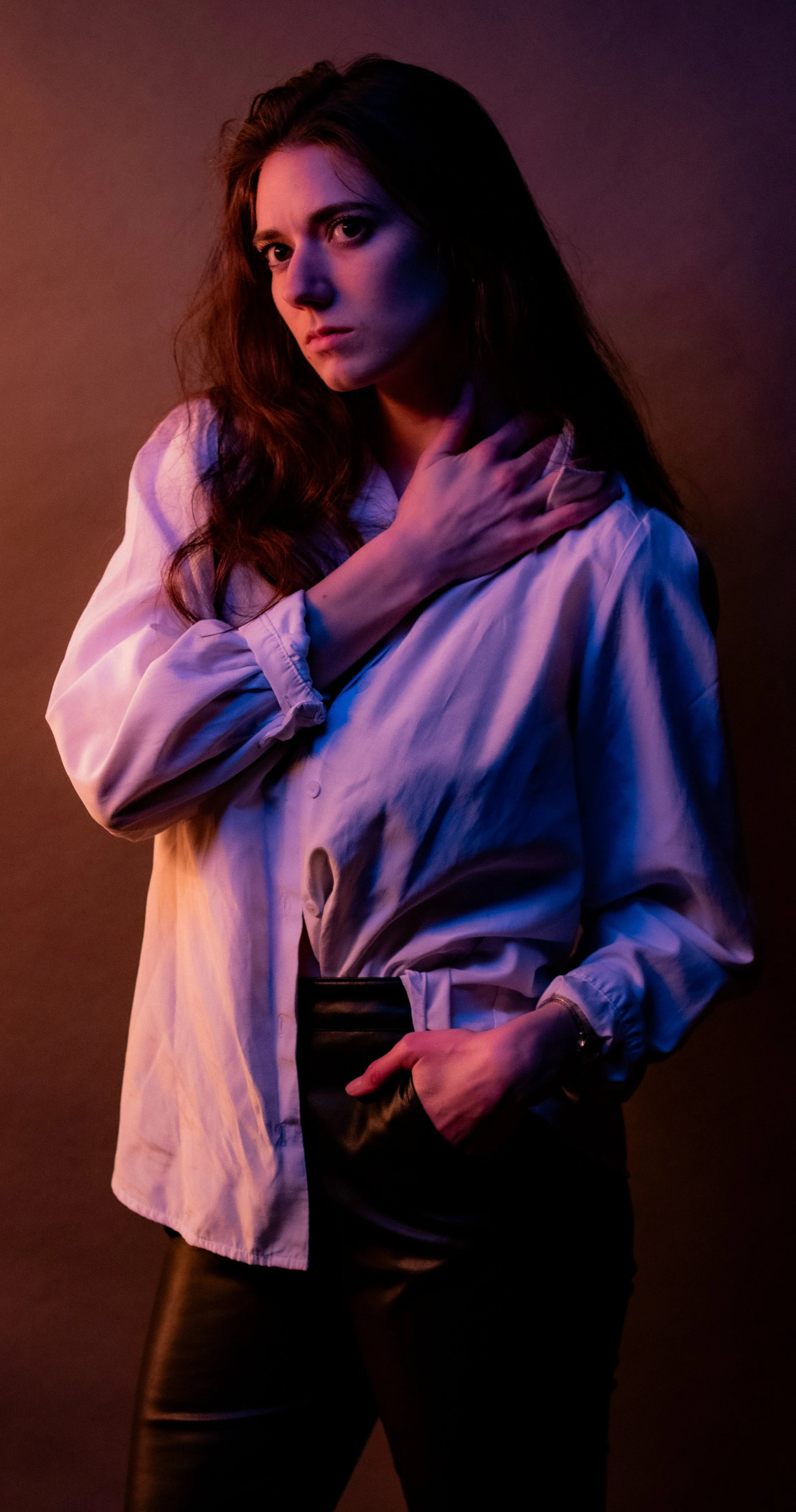A woman with long brown hair poses against a dark background with purple and orange lighting. She is wearing a white oversized shirt, black leather pants, and a belt. She is touching her neck with her left hand and holding a small clutch or purse with her right hand.