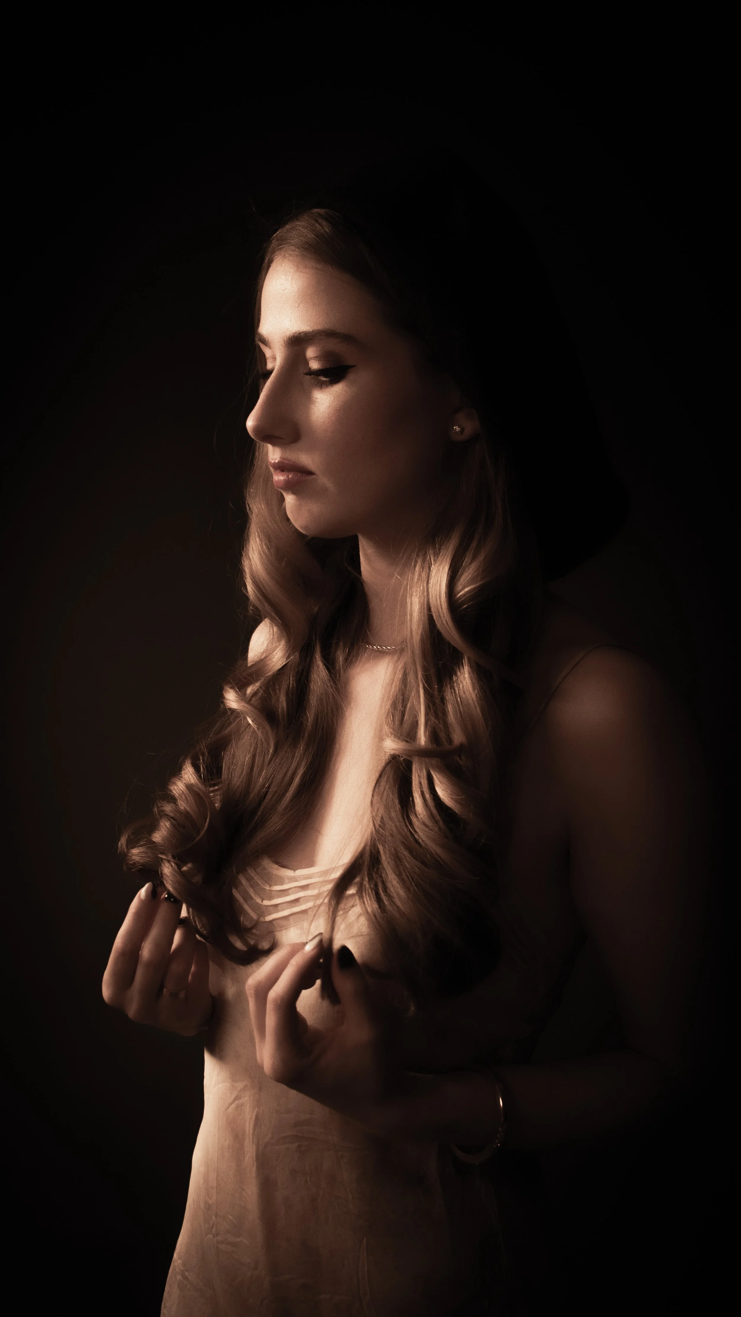 A woman with long wavy hair, closed eyes, and a solemn expression, holding her hands close to her chest, dressed in a light-colored top, against a dark background.