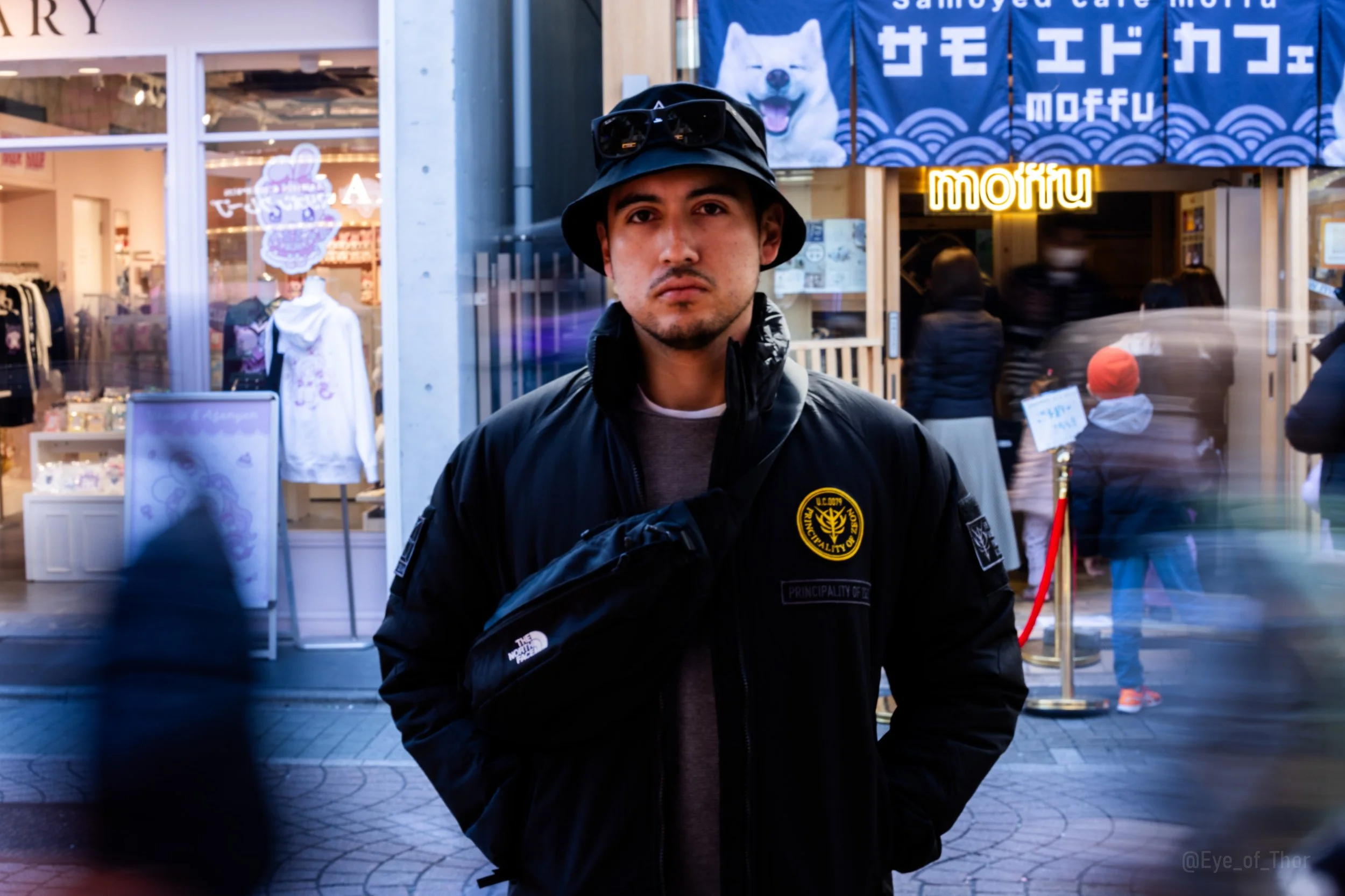 A young man wearing a black bucket hat, sunglasses on his head, and a black jacket standing on a city street in front of storefronts with signs in Japanese and English, including a large blue sign with a white dog image and neon yellow sign reading "moffu".