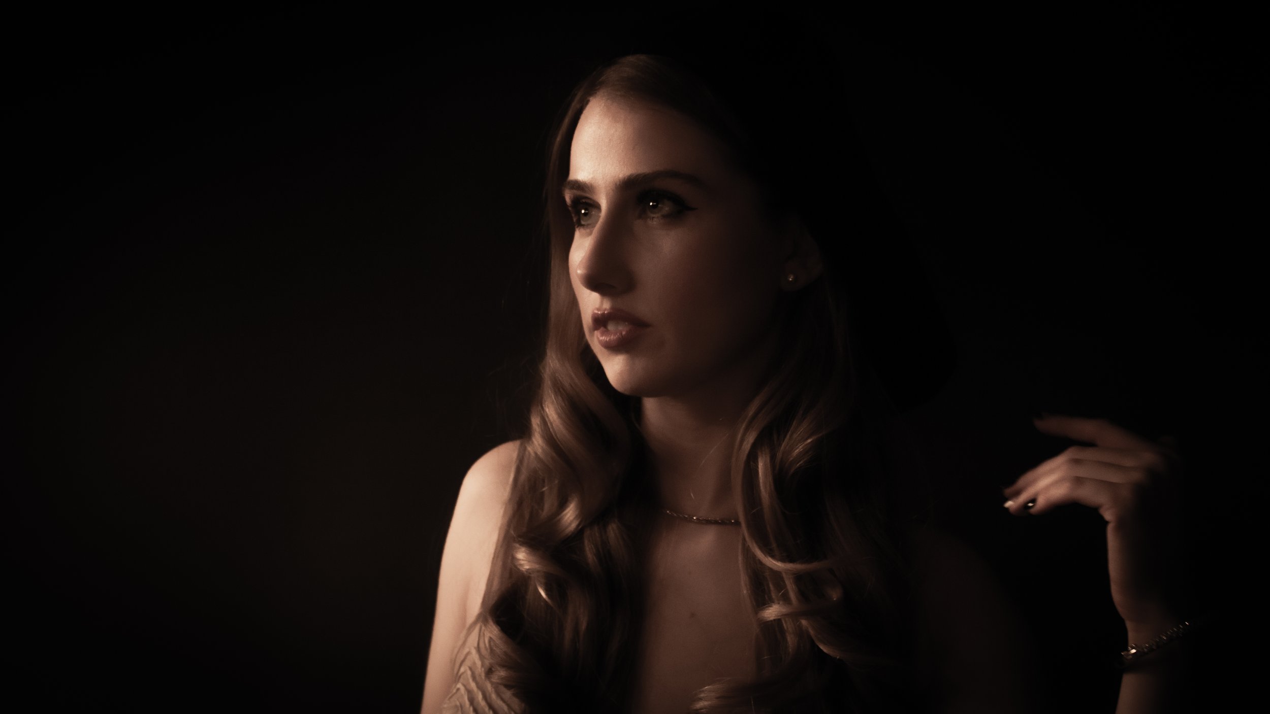 A young woman with long wavy hair, wearing a strapless top, looking to her left, holding her hand near her neck, set against a dark background.