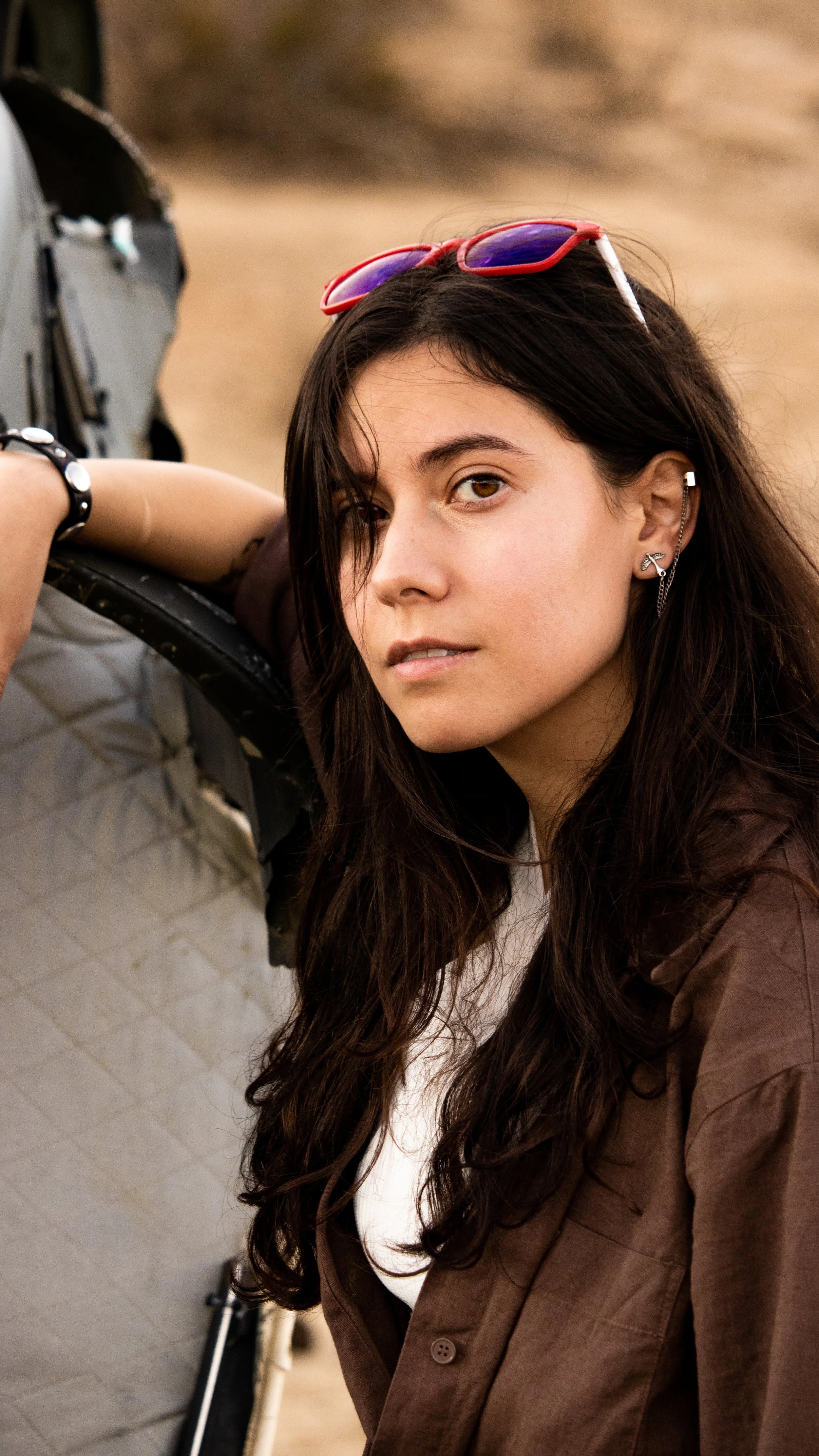 A young woman with long dark hair, wearing sunglasses on her head, earrings, and a brown jacket, leaning on a camera tripod.