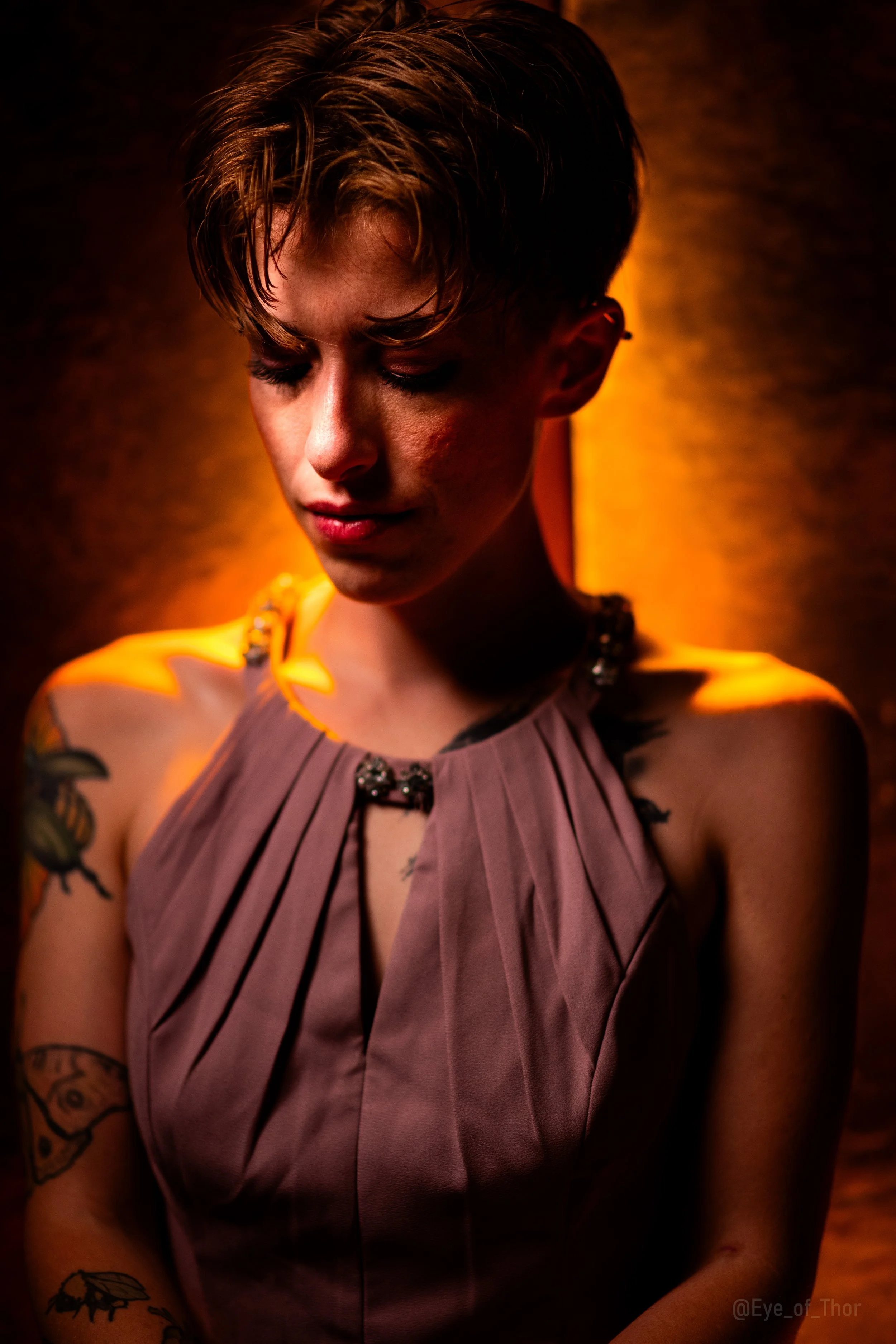A woman with short hair and tattoos on her arm, wearing a mauve pleated dress with a decorative neckline, is looking down against a dark, warm-toned background.