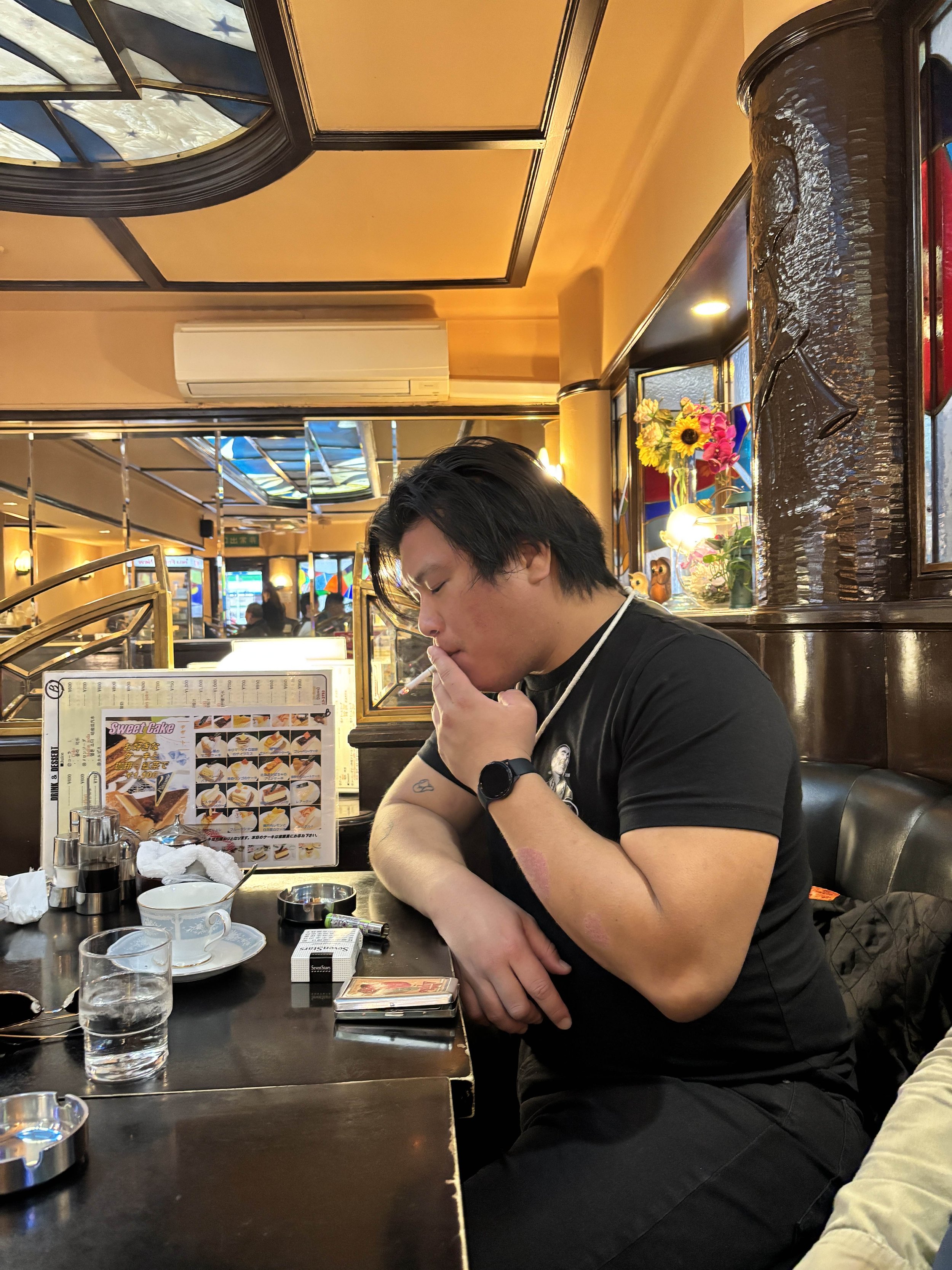 A young man with black hair, wearing a black t-shirt and a black wristwatch, sitting at a restaurant table, appears to be smoking or vaping. The table has glasses, a pack of cigarettes, and a menu with images of desserts. The restaurant has warm lighting, decorative flowers, and stained glass windows in the background.