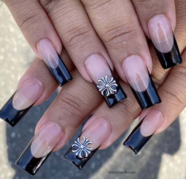 Close-up of hands with acrylic nails featuring French tips with black accents and silver floral embellishments on the middle and ring fingers.
