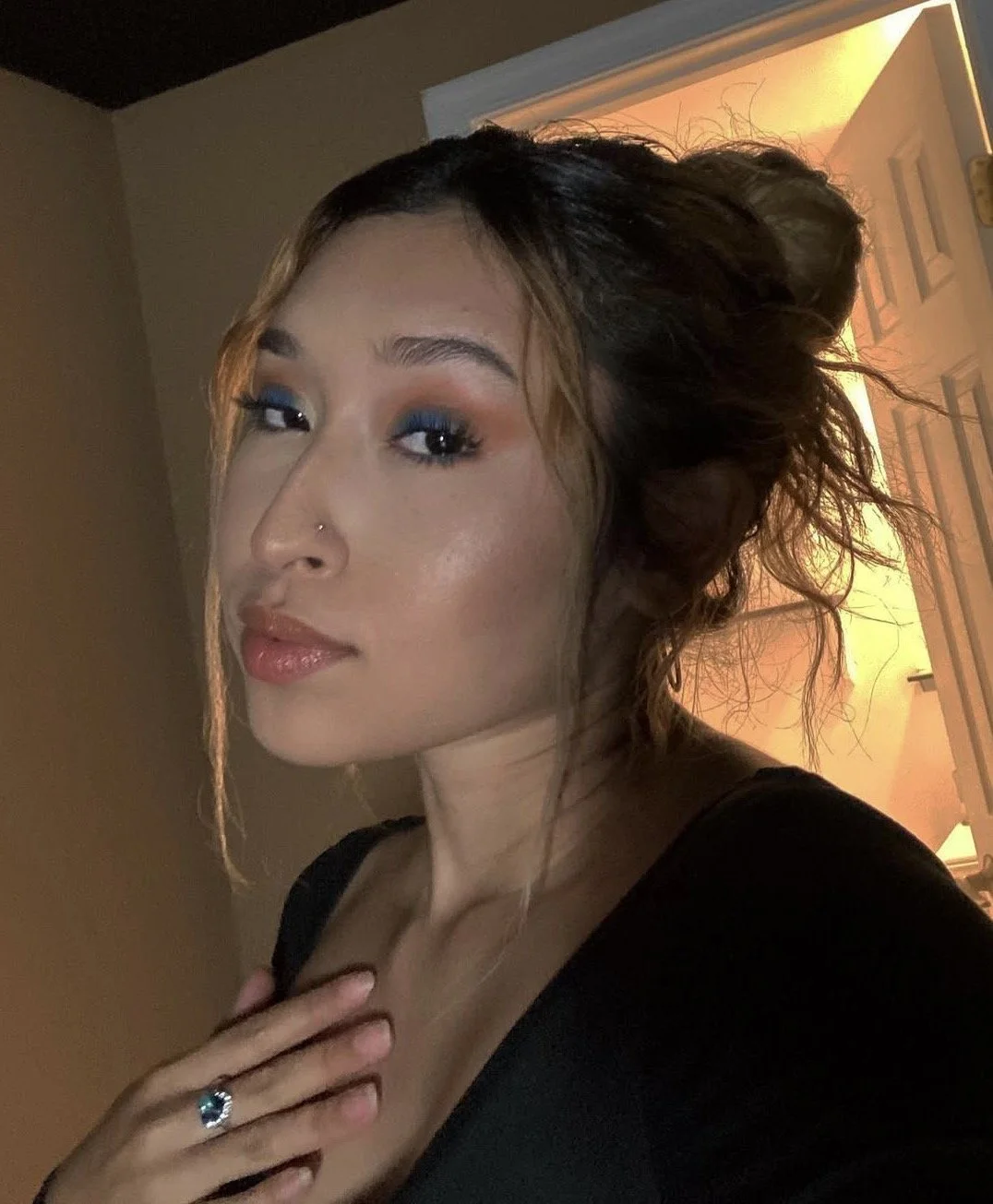 A woman with curly hair styled in an updo, wearing makeup with blue eyeshadow, glossy lips, and a nose piercing, posing indoors with a warmly lit background.