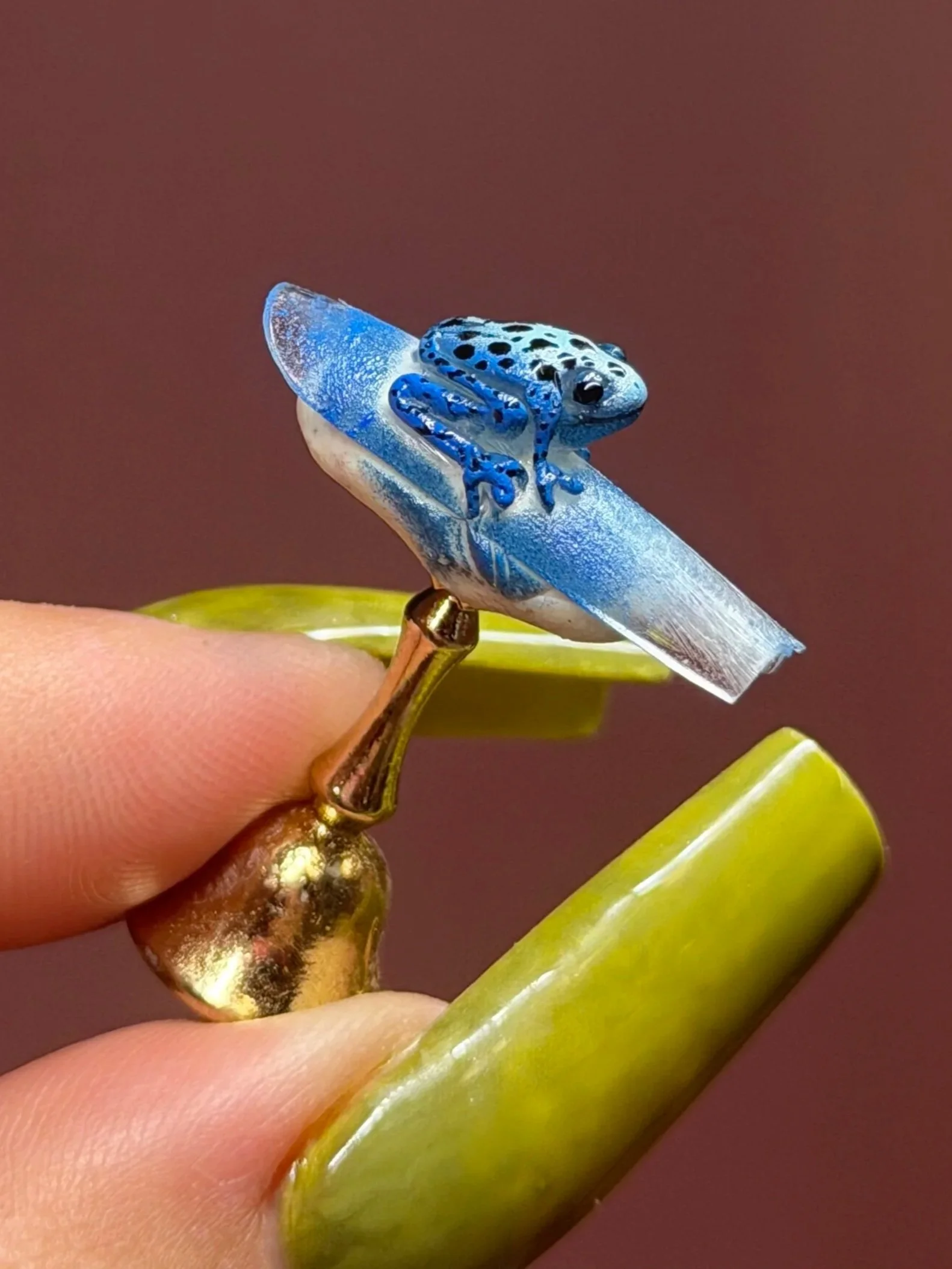 Close-up of a ring with a small, blue frog figurine sitting on a white base, held between a person's fingers with green nail polish.