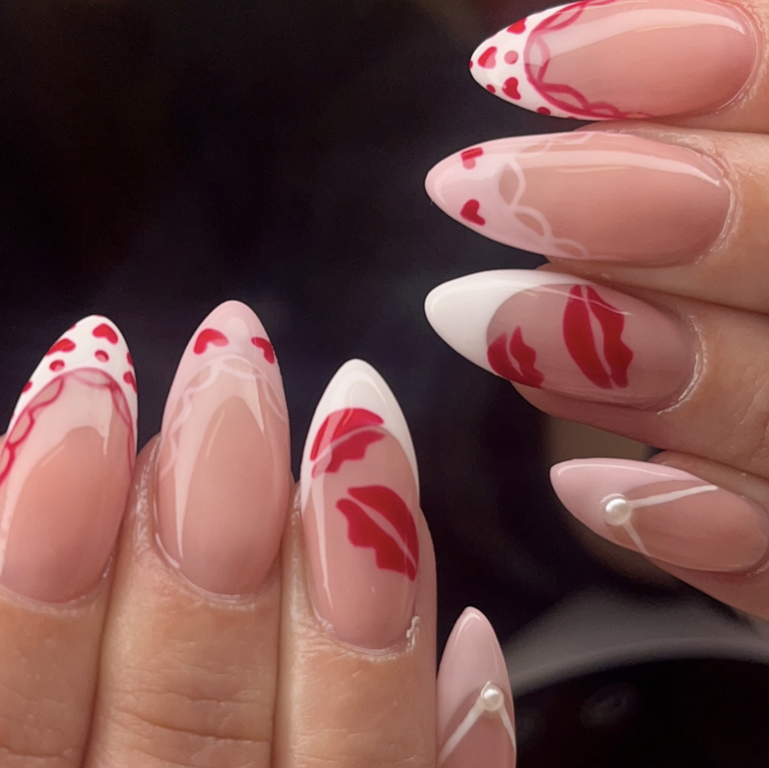 Close-up of decorated almond-shaped nails with pink, white, and red designs, including hearts, lips, and polka dots, set against a black background.