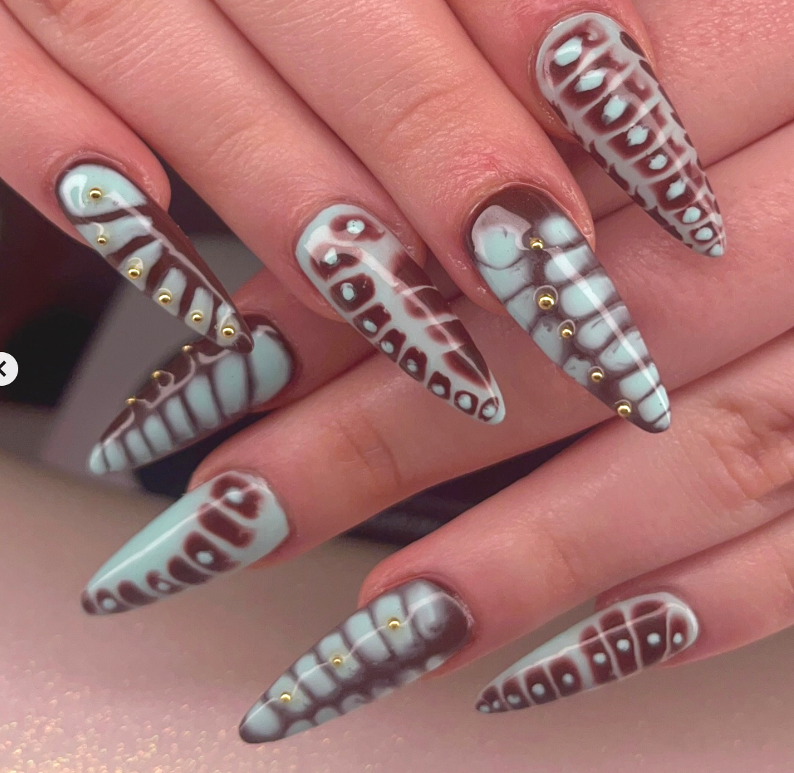 Close-up of long stiletto nails with intricate black, white, and brown geometric leopard print designs, decorated with small gold studs.