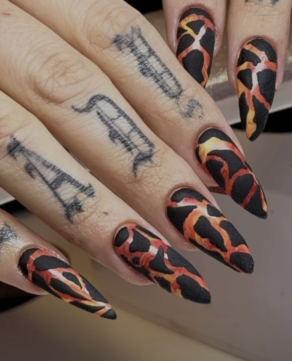 Close-up of a hand with long, sharp, pointy nails painted with a fiery leopard print pattern in red, orange, and black colors. The person has tattoos on their fingers.