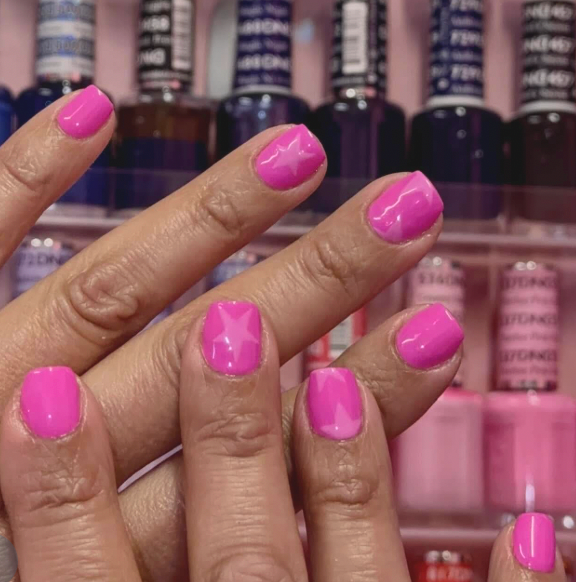 Close-up of a person's fingernails painted in bright pink nail polish, with a nail salon display of various nail polish bottles in the background.