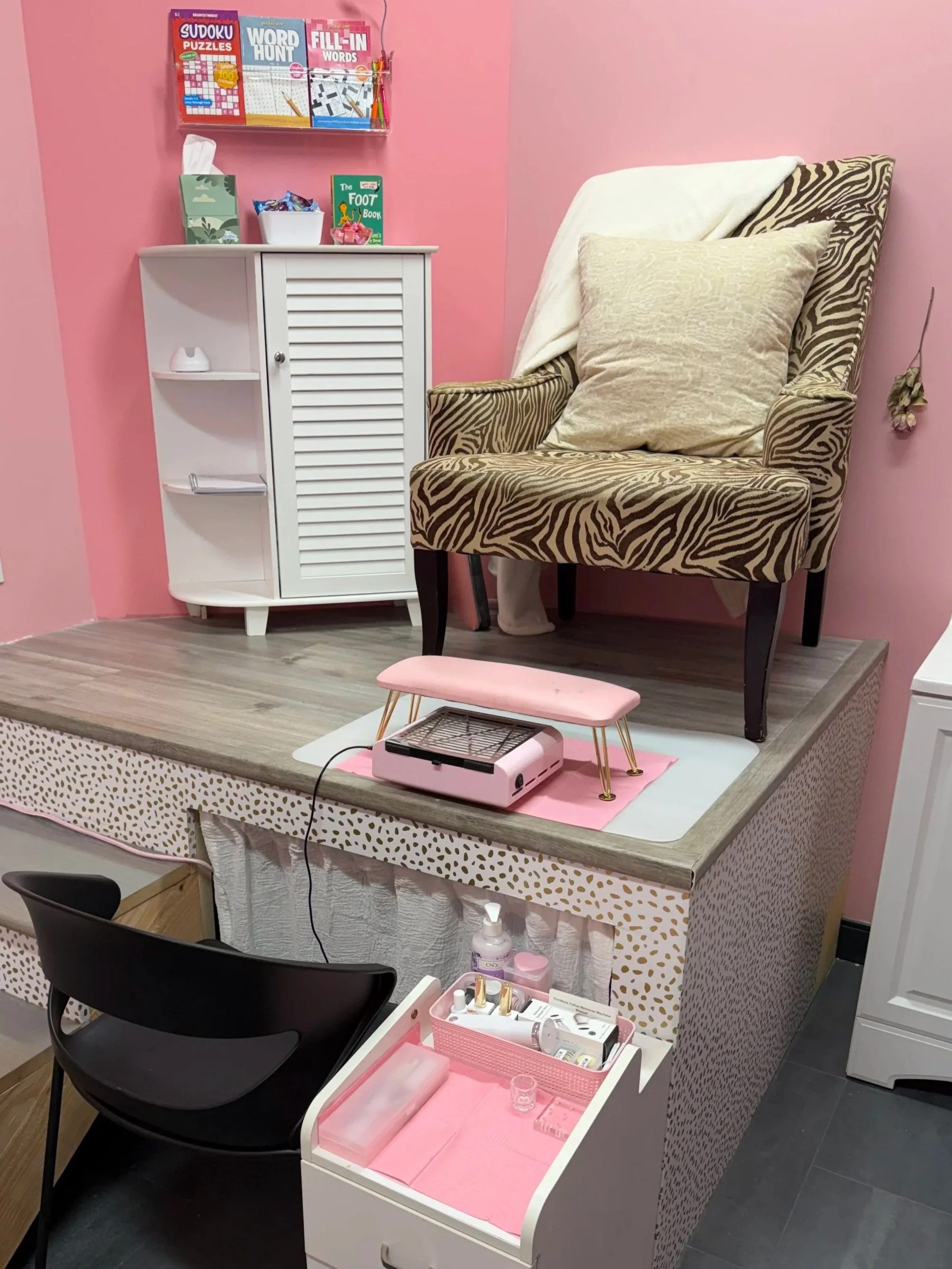 A manicure or beauty salon station with a pink theme. It has a raised platform with nail care supplies, a black chair, a small white trolley with nail polish tools, a pink nail dryer, and a black chair. The back wall features pink walls, a white cabinet, and decorative items. A shelf above holds books and stationery, and a small pink magnifying lamp is on a table.