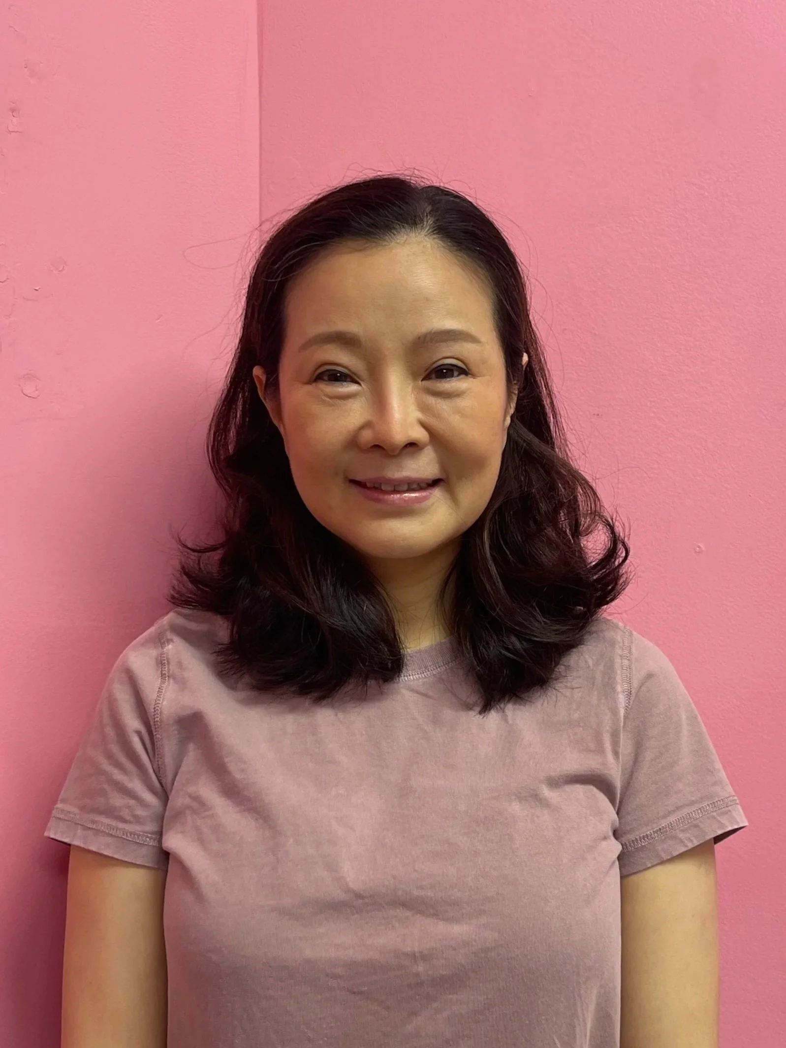 A woman with shoulder-length wavy dark hair smiling at the camera, standing in front of a pink wall.