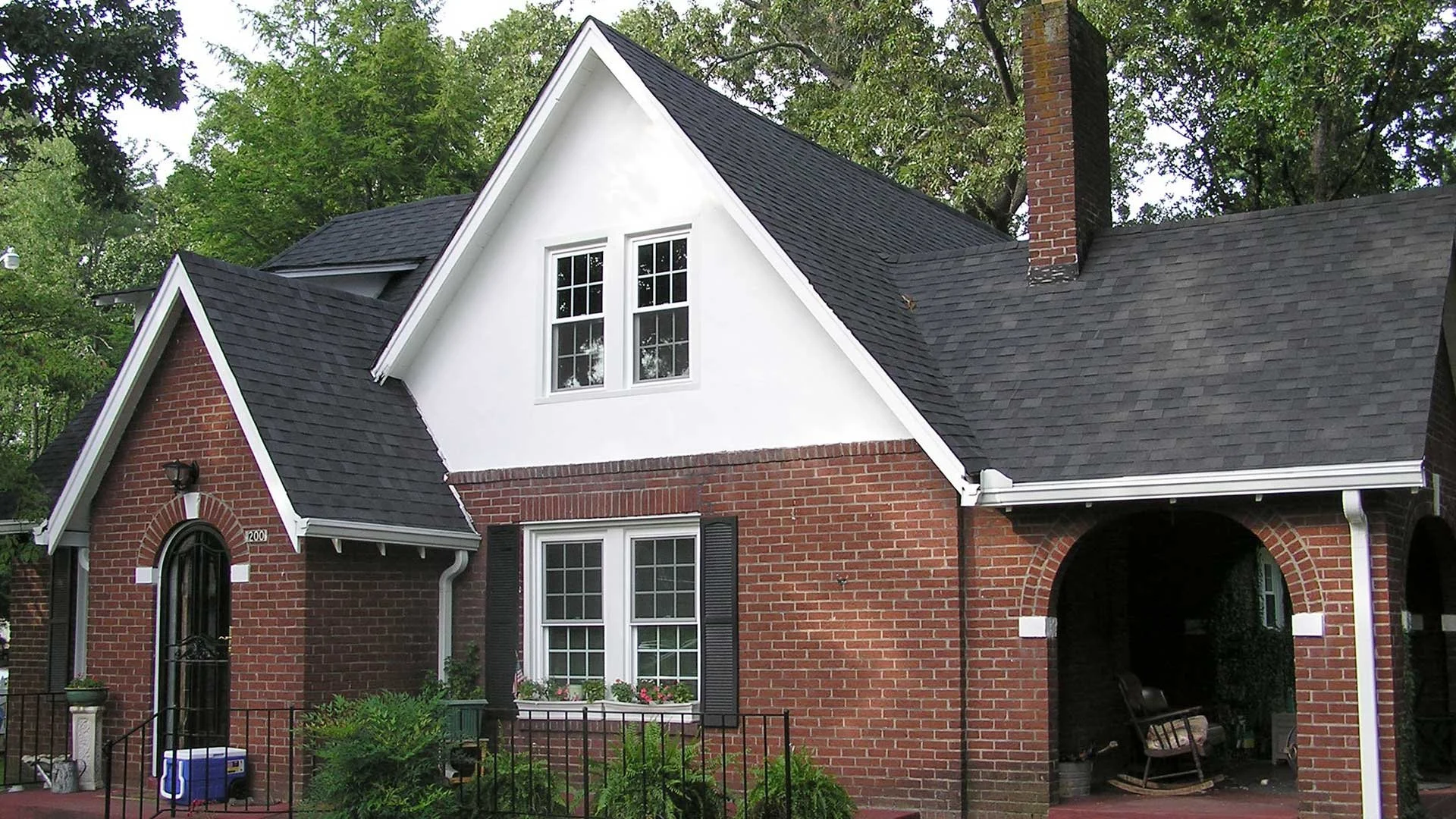 A two-story brick house with black shutters, white trim, and a steep black shingle roof, surrounded by trees and a small front yard.