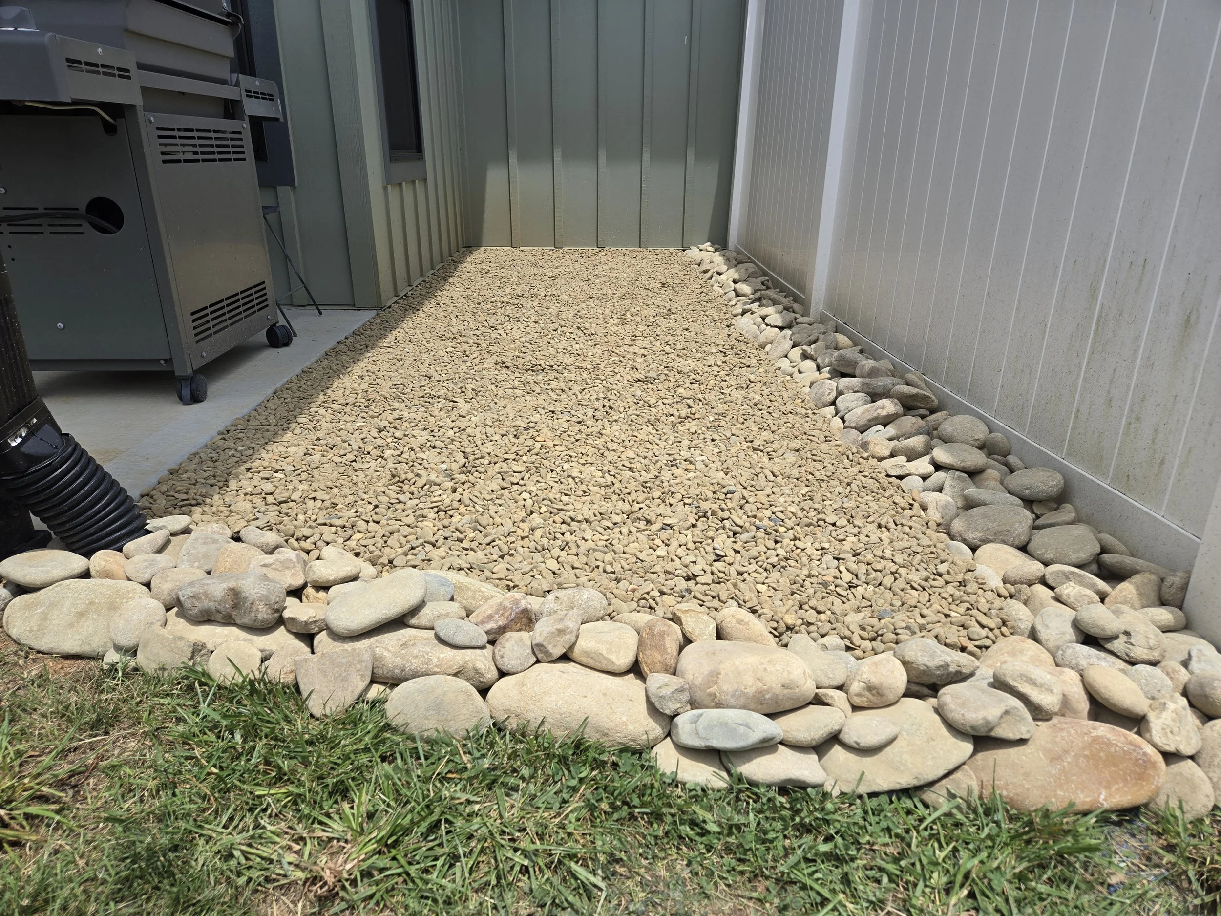 A backyard area with gravel ground, bordered by a row of large smooth rocks. On one side, there are some patches of grass and on the other, a gray metal fence or wall. There is a vacuum or large hose in the bottom left corner and some metal equipment on the left side of the image.