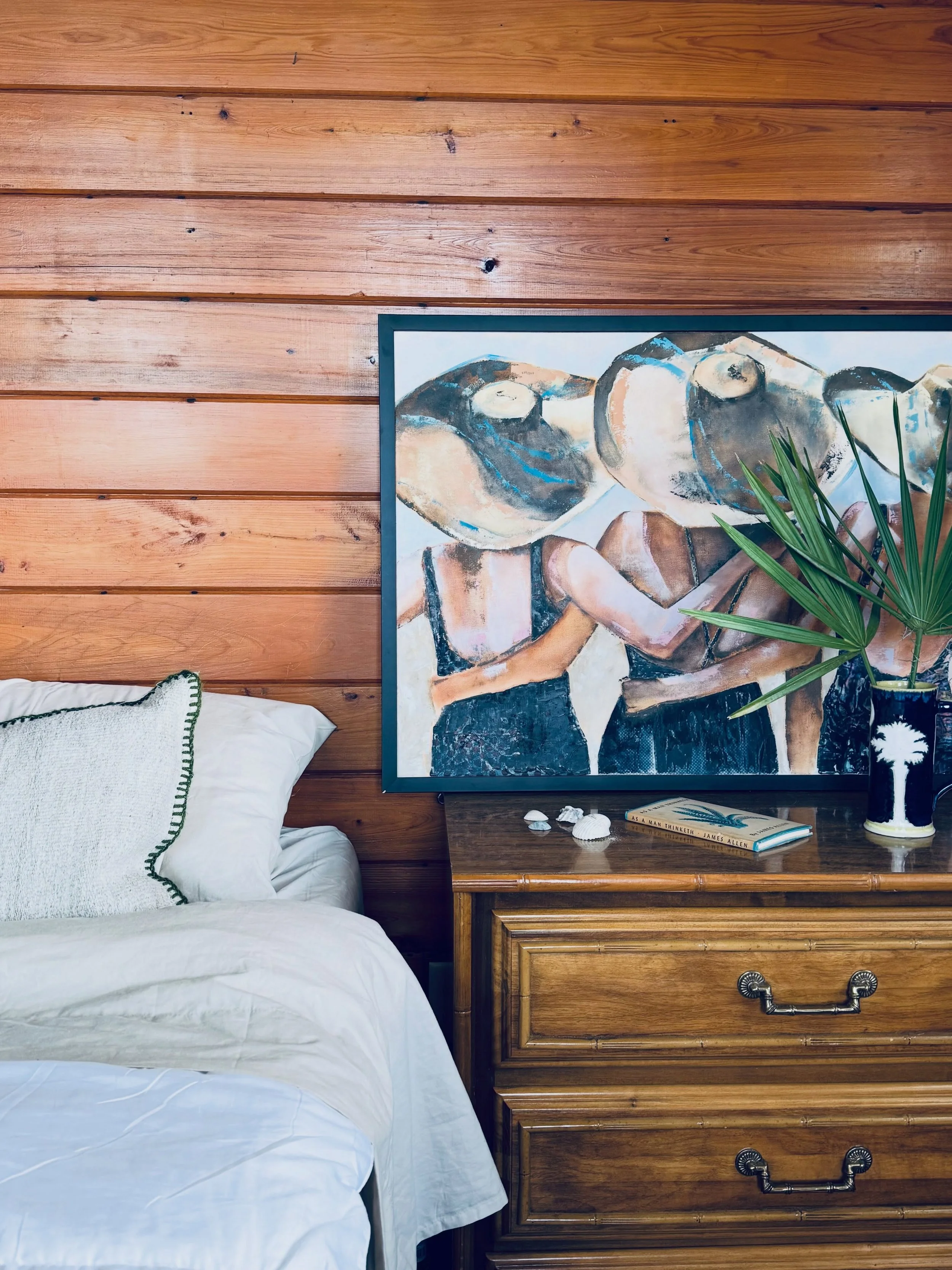 A cozy bedroom corner with a bed and a wooden nightstand. The bed has white sheets and a pillow with green piping. The nightstand holds a painting of abstract figures, a few seashells, a book, and a potted plant with long green leaves. The wall behind is covered in horizontal wooden planks.