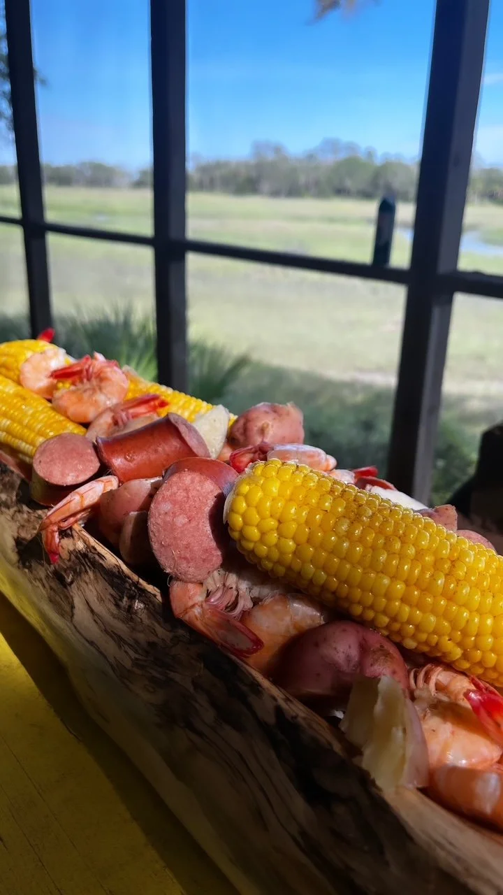 A rustic wooden serving platter with shrimp, sliced sausage, boiled corn on the cob, and potatoes, set against a backdrop of a screened porch with a grassy landscape and trees in the distance.