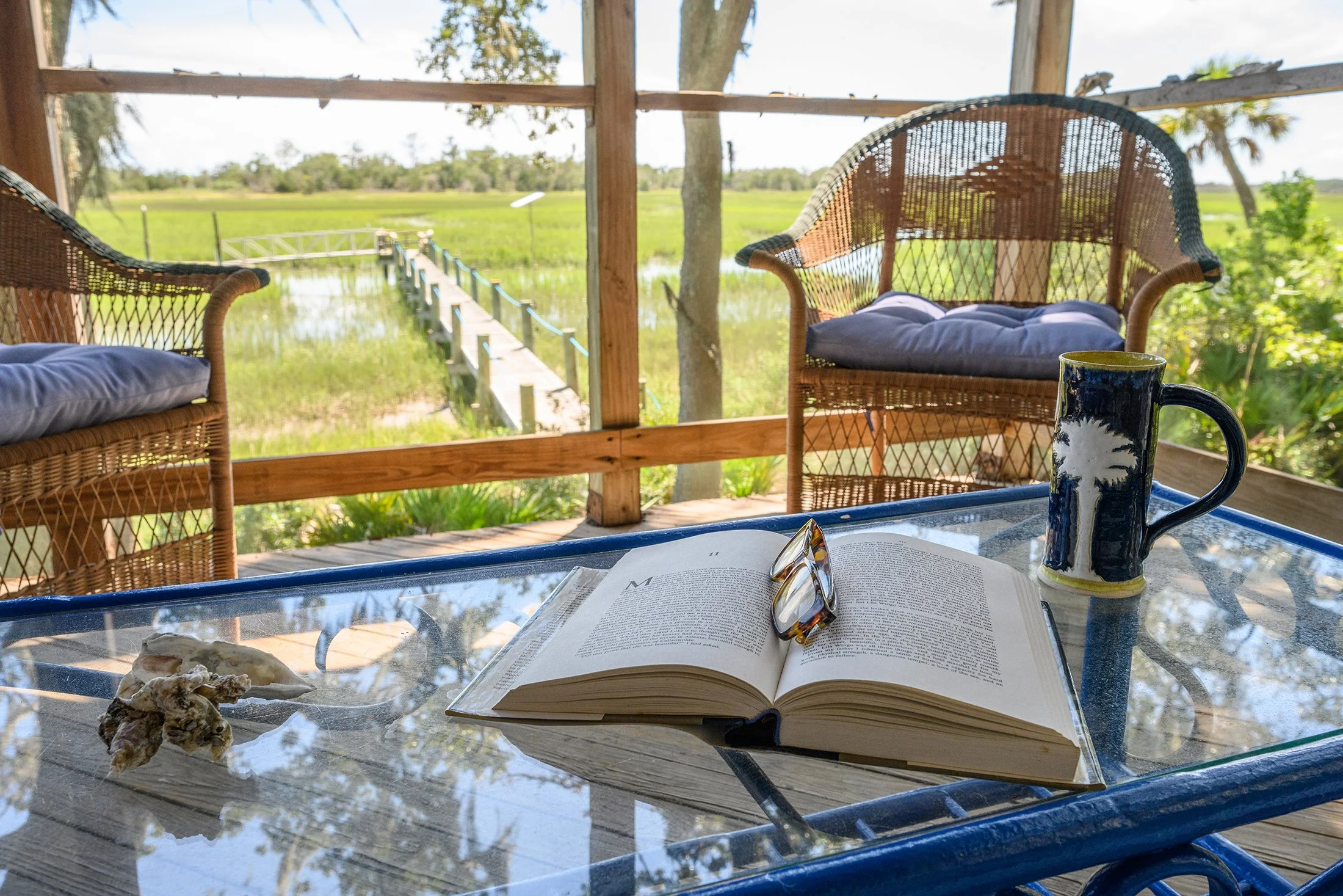 A table with an open book, reading glasses, and a mug set outdoors on a porch overlooking a marsh with green grass and water, with wicker chairs on the porch and trees in the background.