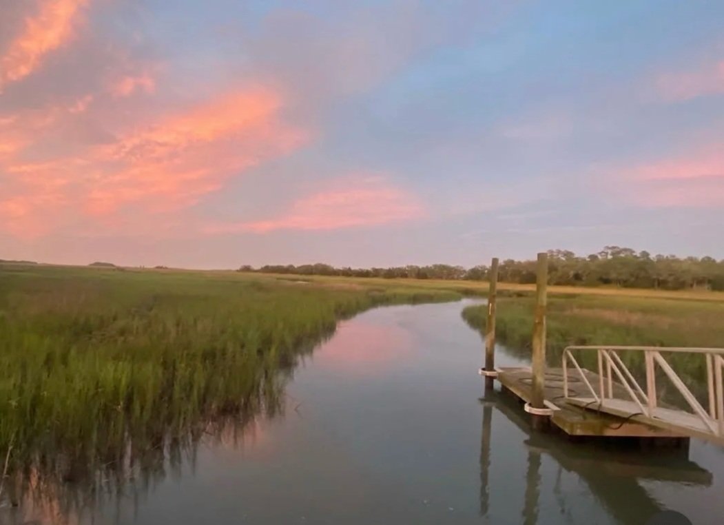 A peaceful marshland scene during sunset with a calm waterway, green marsh grass, and a wooden dock extending into the water, under a sky with pink and blue clouds.