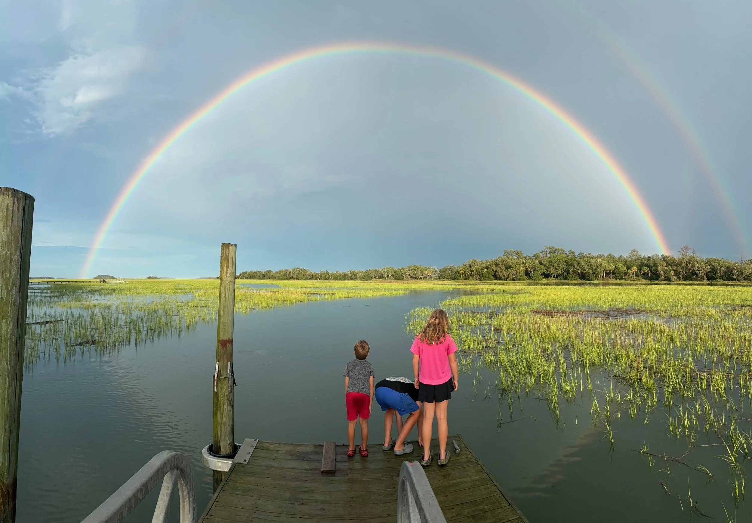Three children stand on a small wooden dock by a pond, watching a rainbow in a partly cloudy sky over a green landscape.