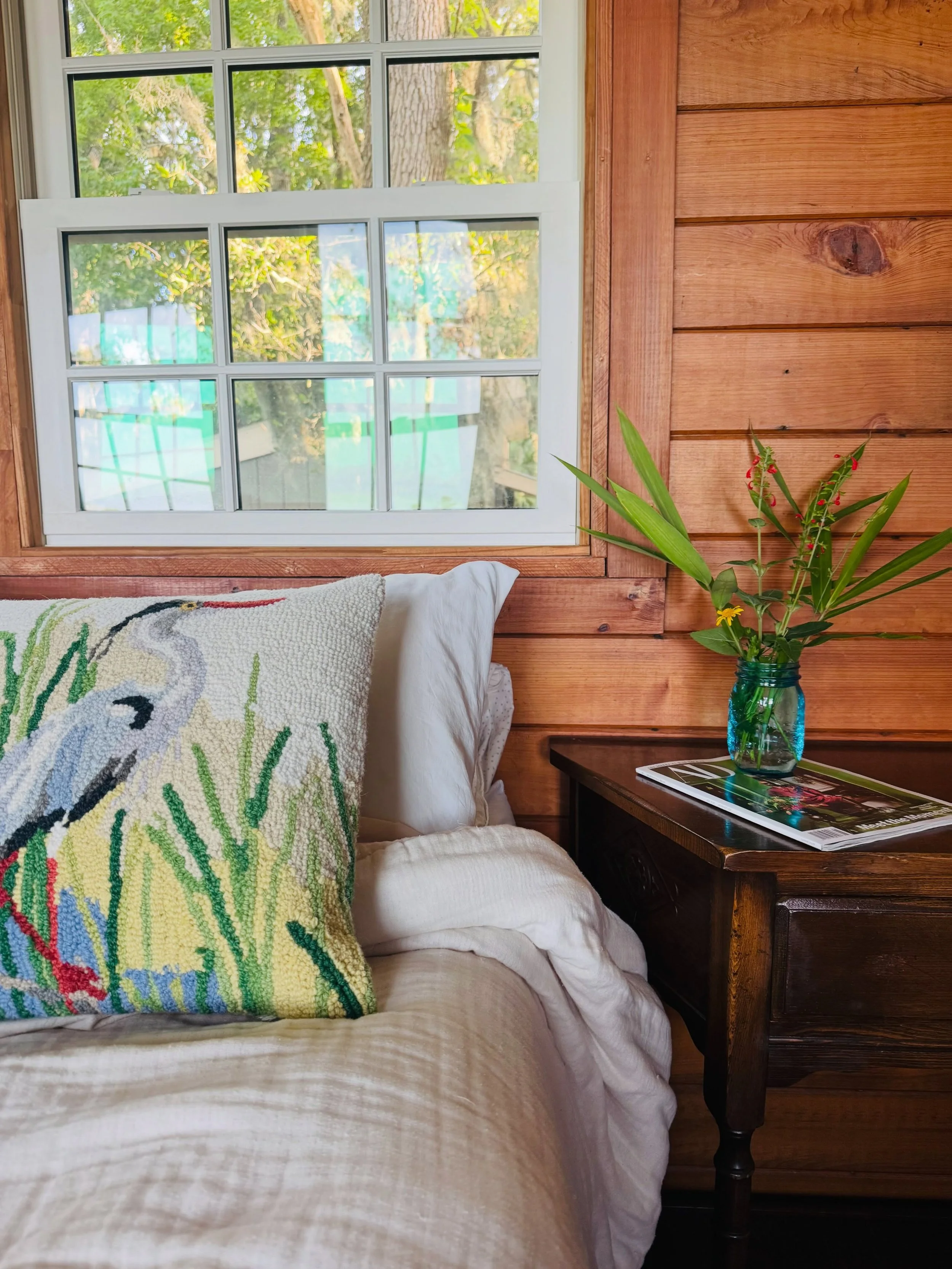 Close-up of a cozy interior corner featuring a white bed with a decorative pillow, a wooden nightstand with a vase of green plants, and a window showing green outdoor foliage.