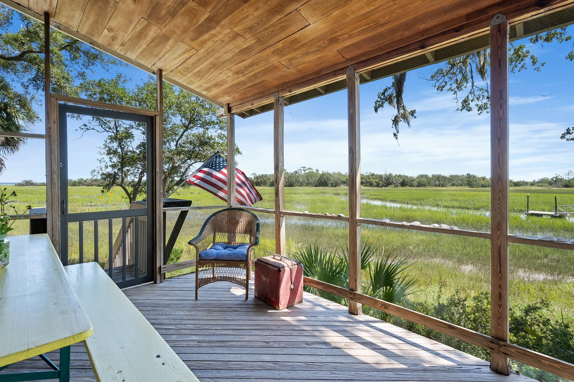 View of an open porch with wooden ceiling and railings overlooking a grassy wetland area with trees under a blue sky. There is a wicker chair with a blue cushion, a small table, an American flag, and a red cooler on the porch.