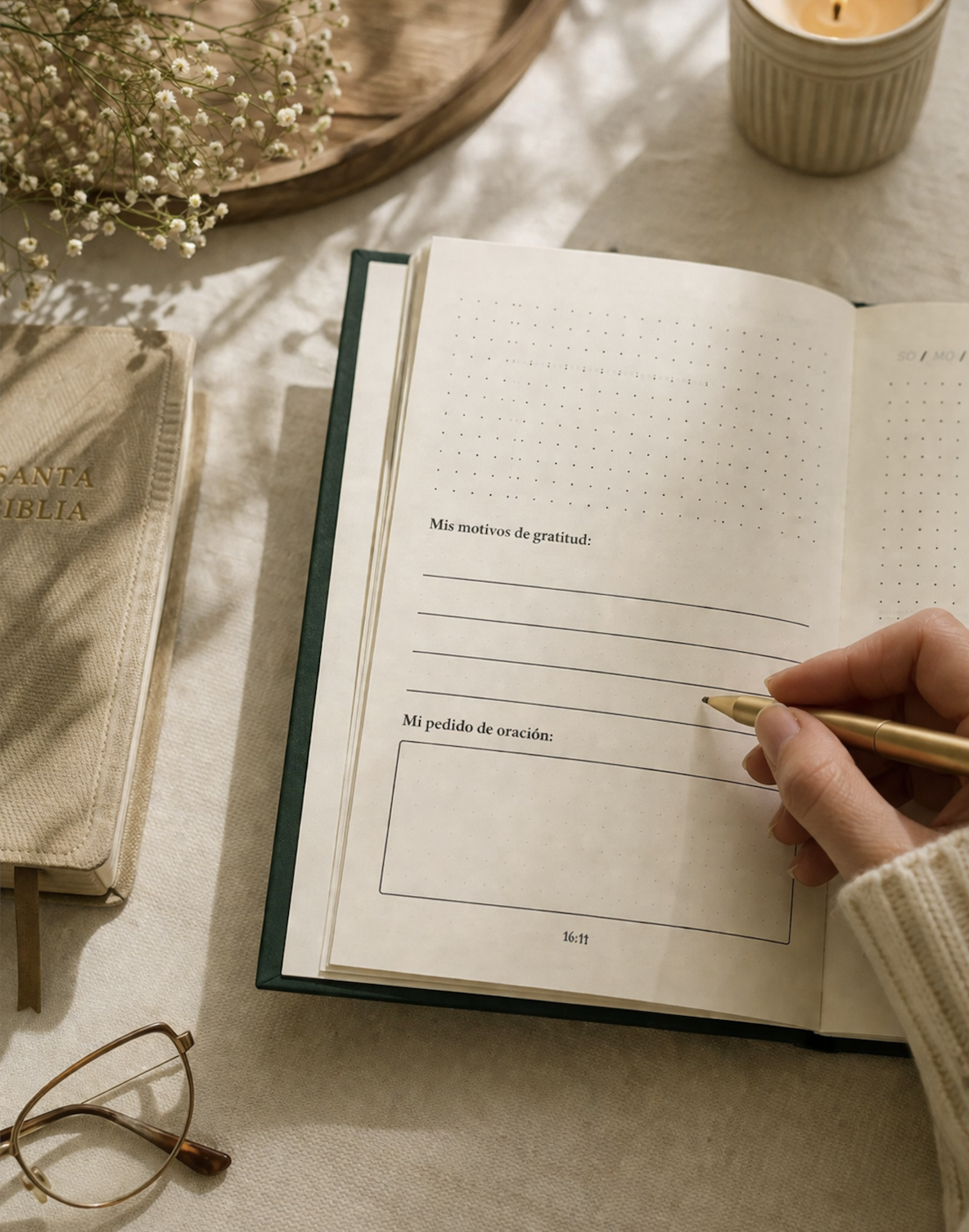 Libreta de notas abierta en una mesa, con una mano escribiendo con bolígrafo dorado en ella. En la página hay espacios para escribir motivos de gratitud y una oración. También se ven un libro de la Biblia, unas gafas, una vela y flores en una bandeja.