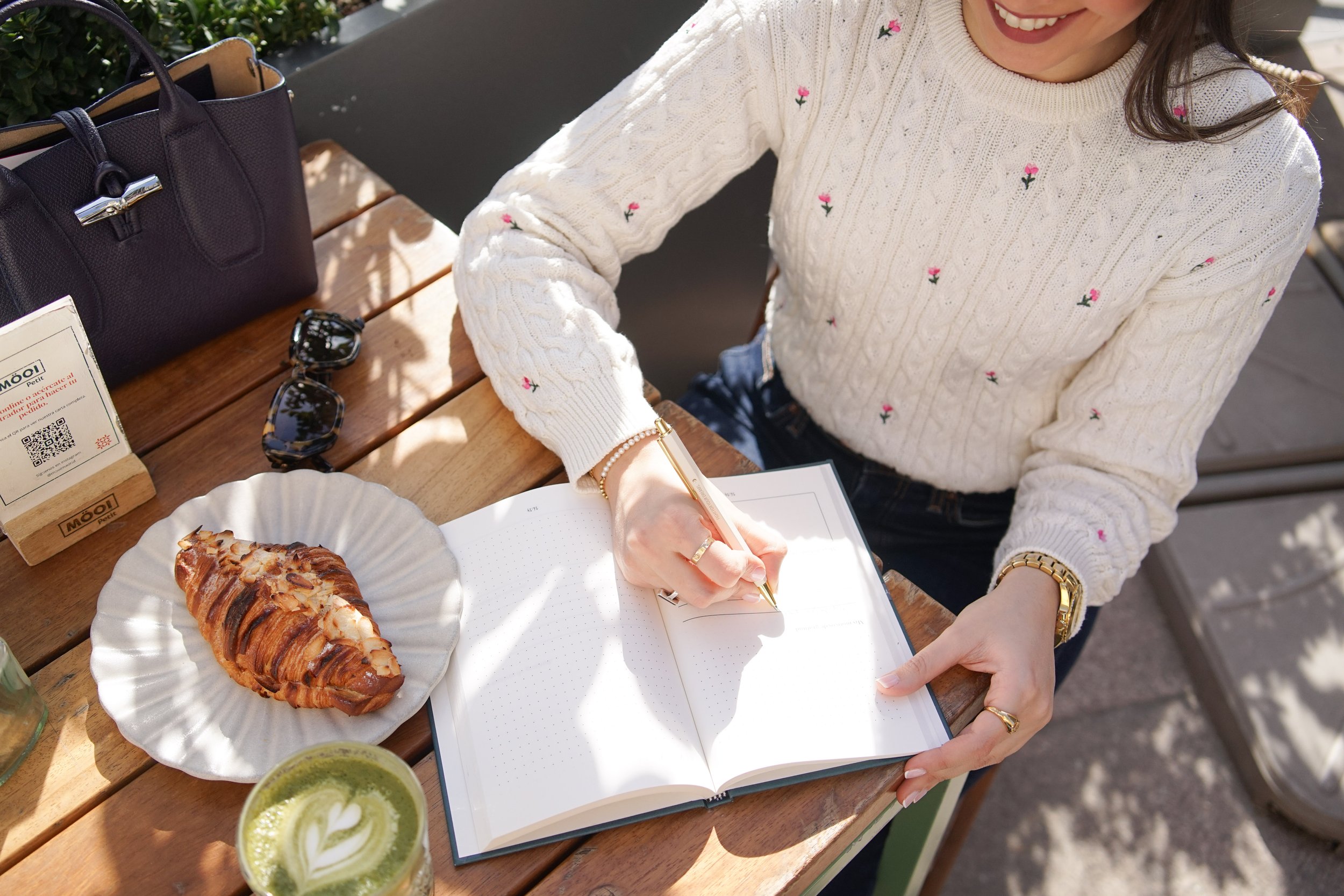 Persona escribiendo en un cuaderno en una mesa de madera con comida y bebidas, incluyendo un croissant, té matcha y un par de gafas.