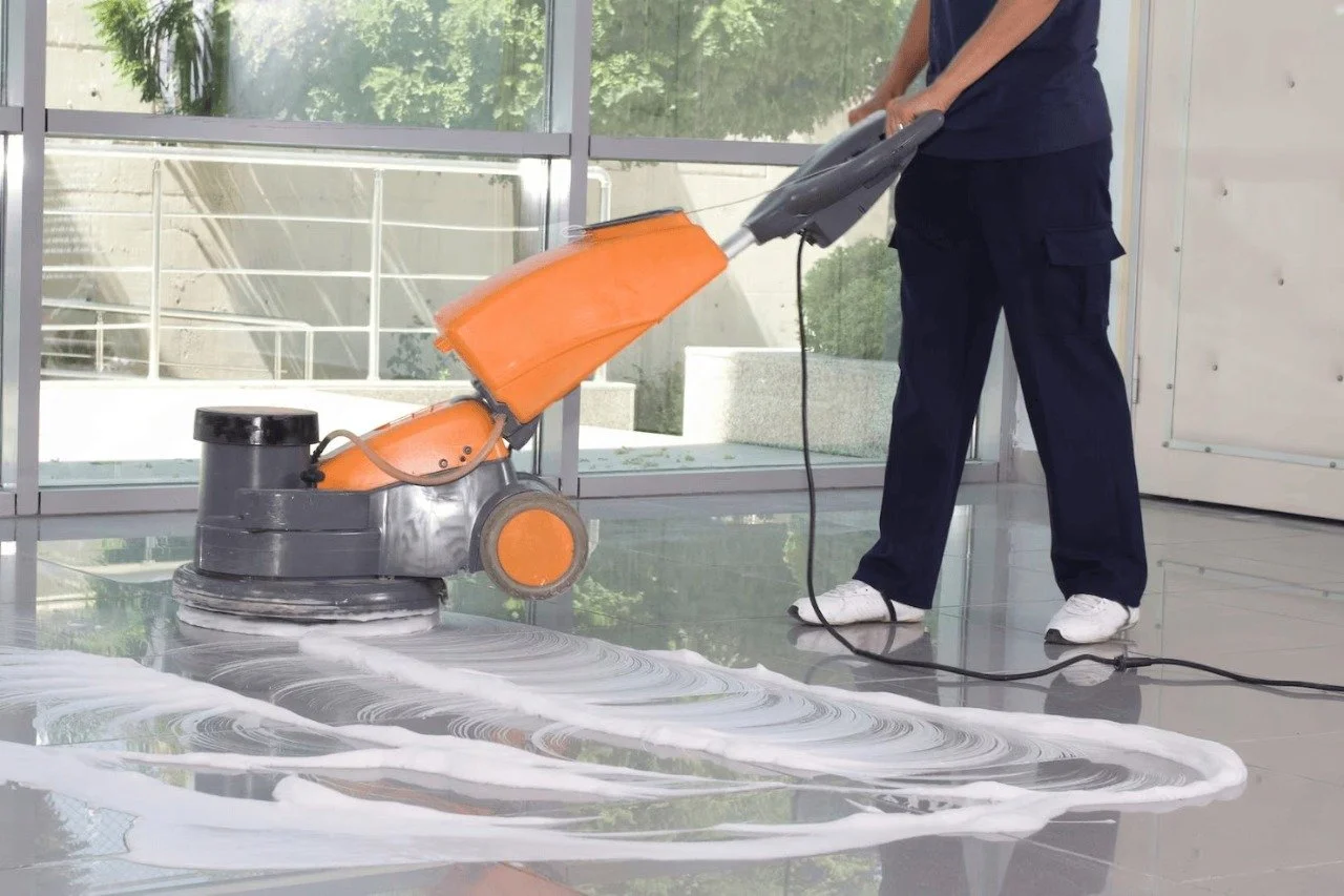 A person cleaning a shiny tile floor with a floor buffer machine, which produces soap or cleaning solution streaks on the floor.