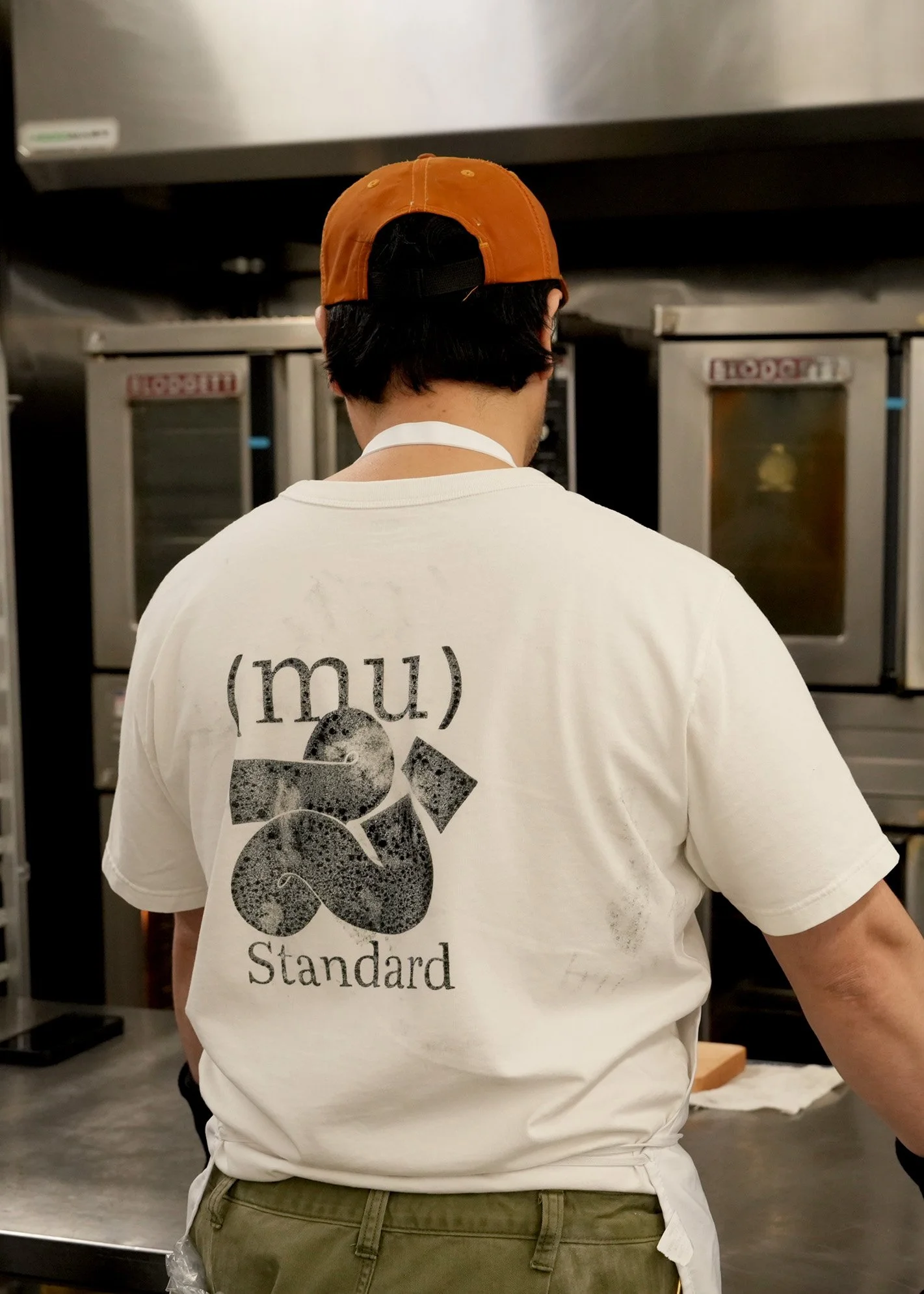 Person with black hair wearing an orange and black cap, a white T-shirt with text and graphics on the back, and olive green pants, standing in front of industrial kitchen equipment.