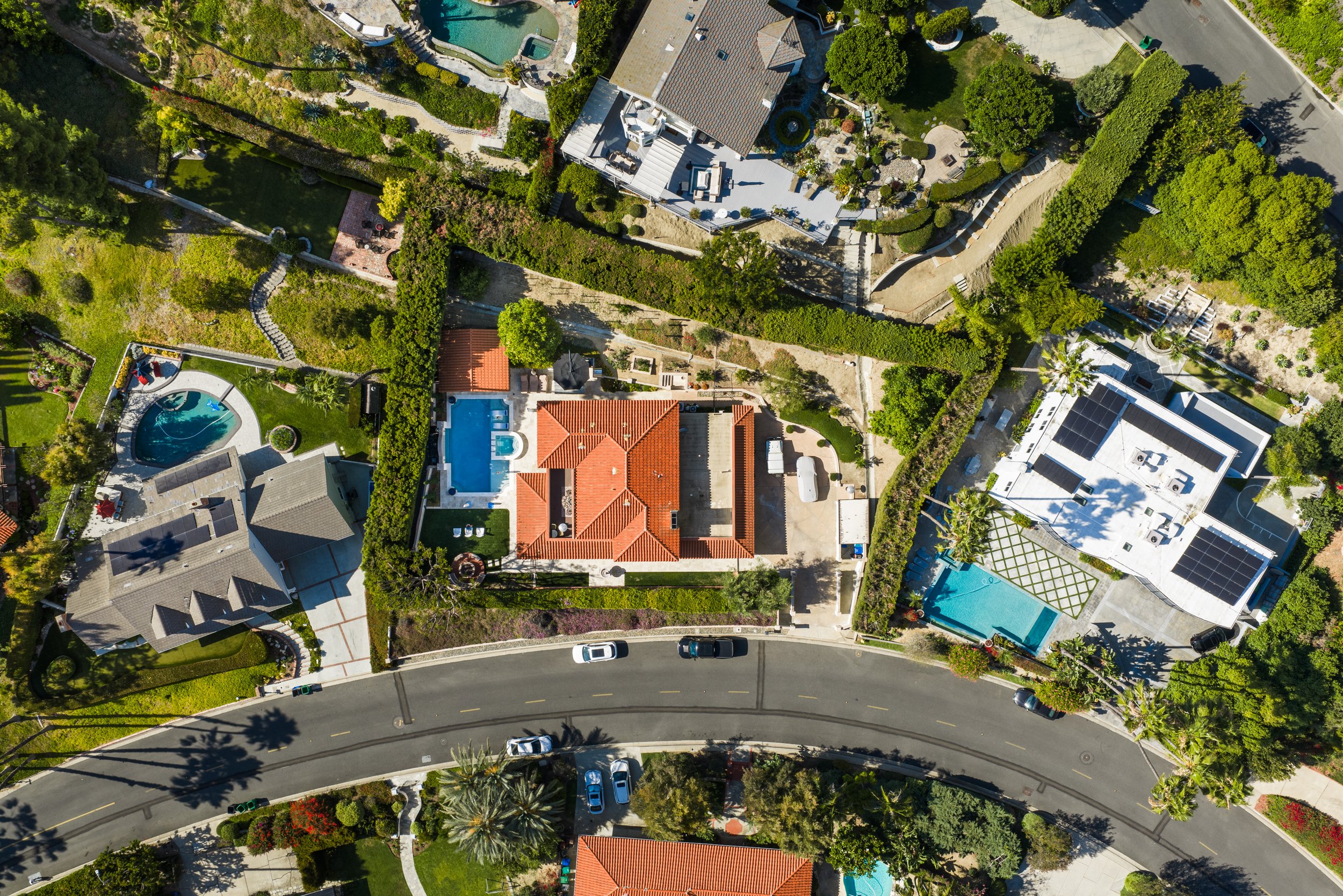An aerial view of a neighborhood with houses surrounded by greenery, trees, and yards. Visible features include swimming pools, a street with parked cars, and landscaped gardens.