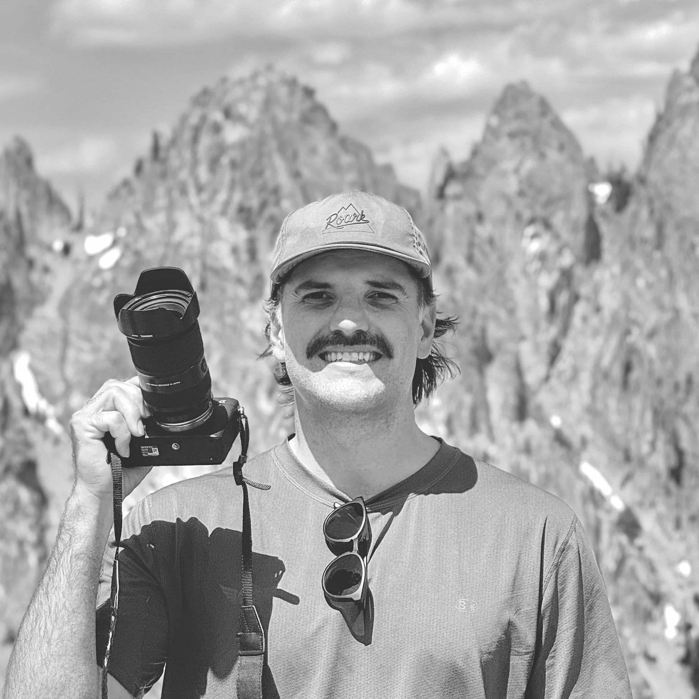 A man with dark hair, mustache, and beard smiling, wearing a baseball cap with the word 'Roar' on it, carrying a backpack, holding a camera, and wearing sunglasses hanging from his shirt. The background features a scenic mountain landscape in black and white.