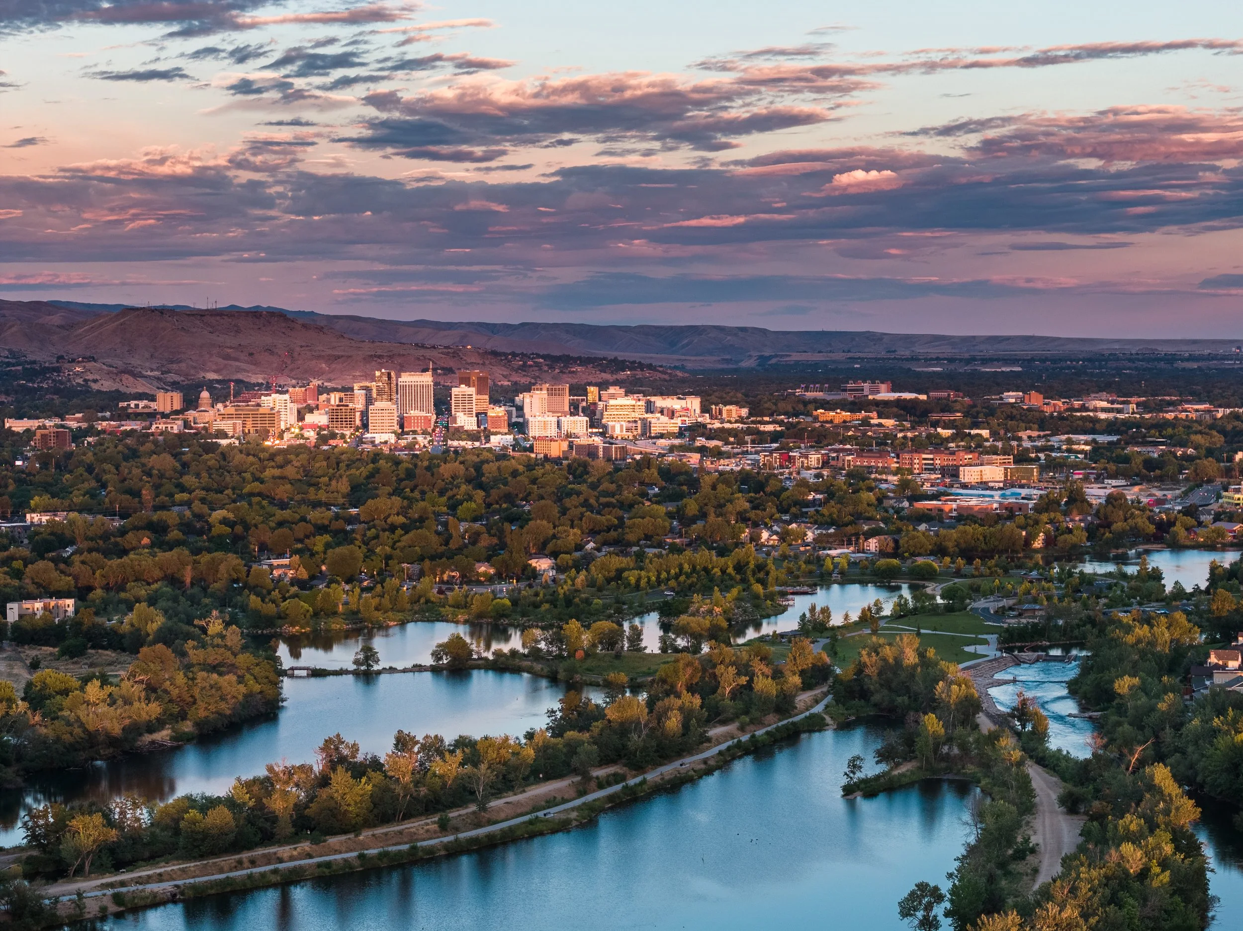Boise city skyline at sunset with tall buildings, surrounded by green trees and a river in the foreground, with mountains in the background and a sky filled with pink and purple clouds.